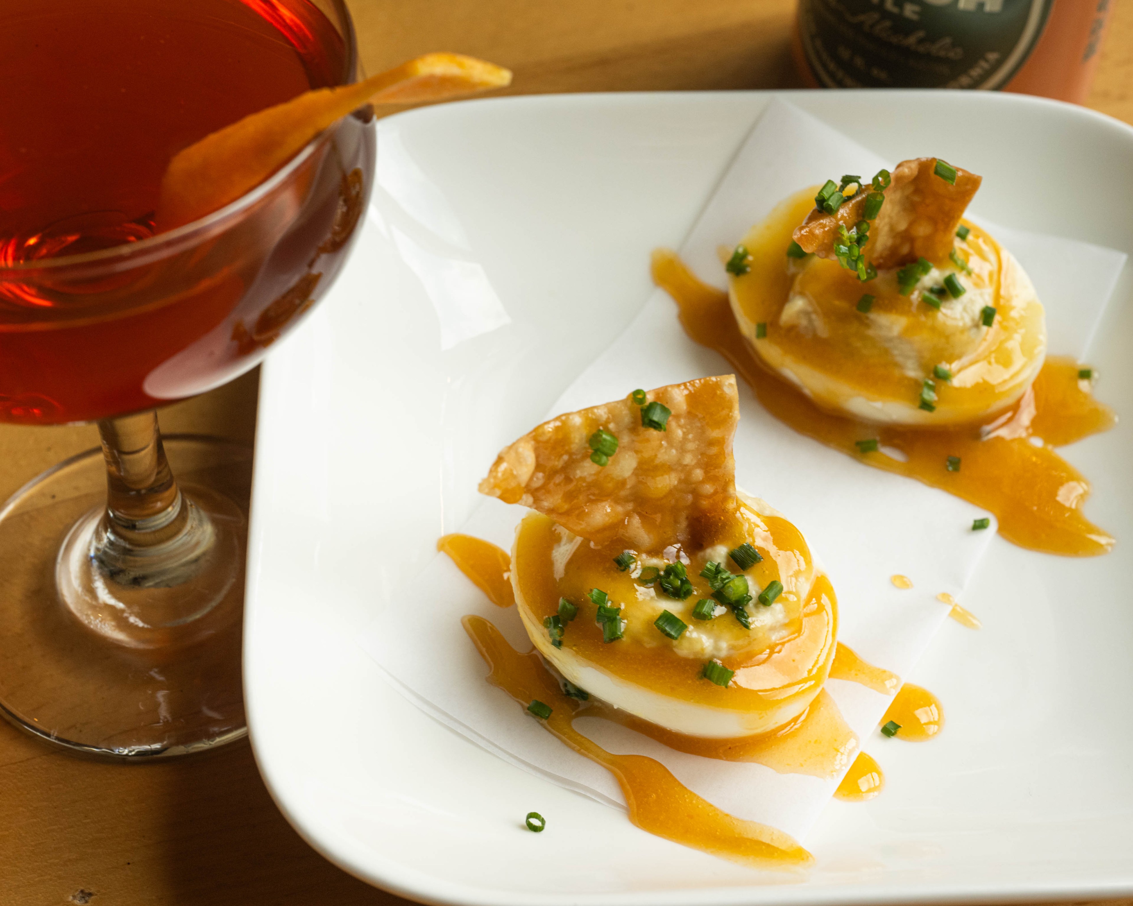 A cocktail with an orange peel beside a white plate holding two deviled eggs topped with sauce, chives, and crispy toppings, on a wooden table.