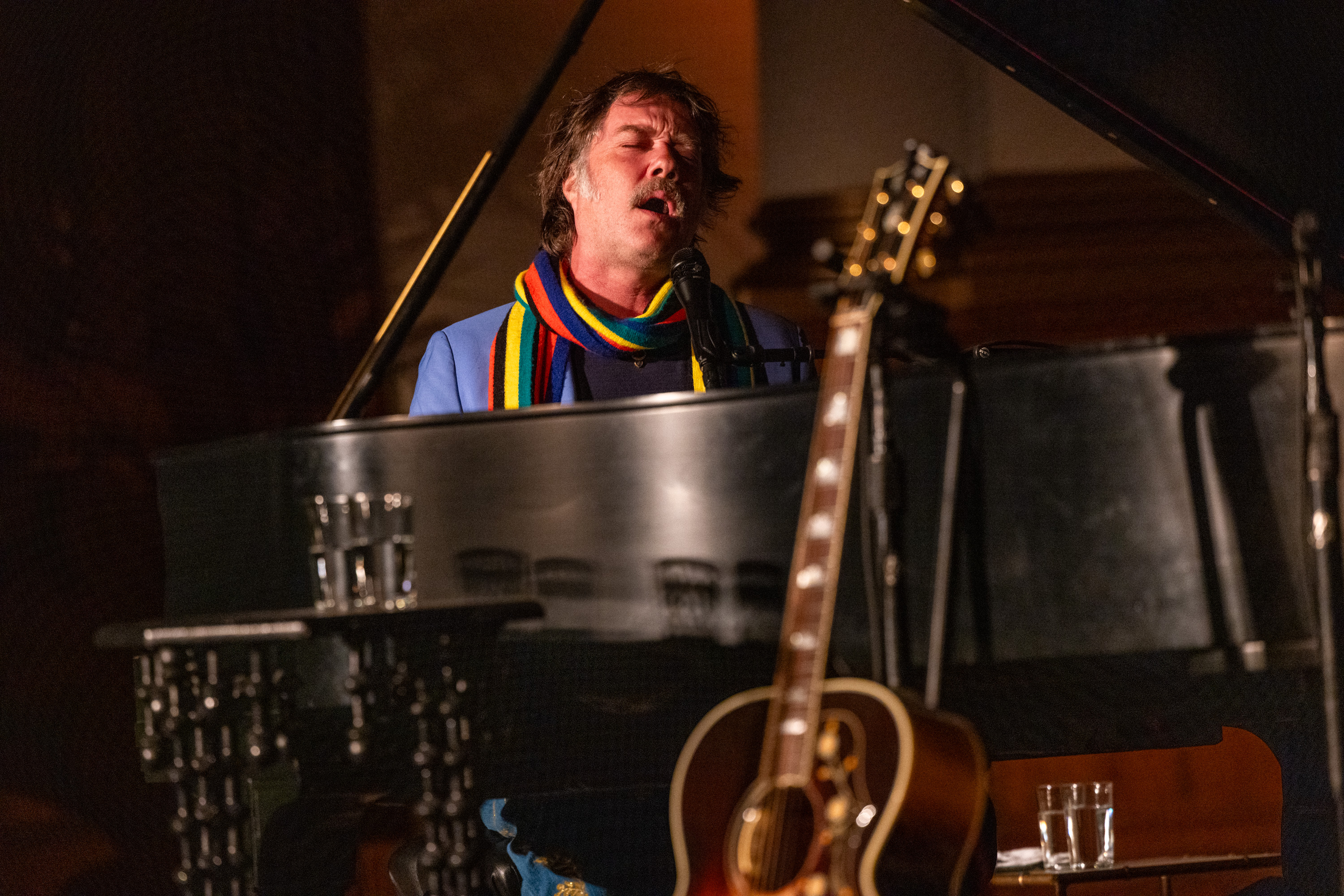 A person passionately sings at a black grand piano, wearing a colorful striped scarf. A guitar leans against the piano, and there are glasses of water nearby.