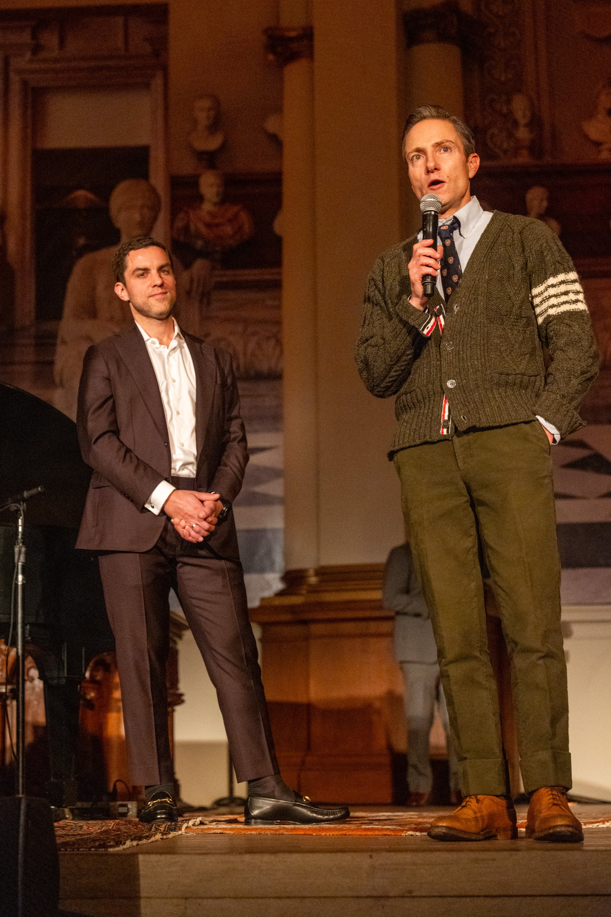 Two men stand on a stage. One is speaking into a microphone wearing a green cardigan and khaki pants; the other in a dark suit stands listening attentively.