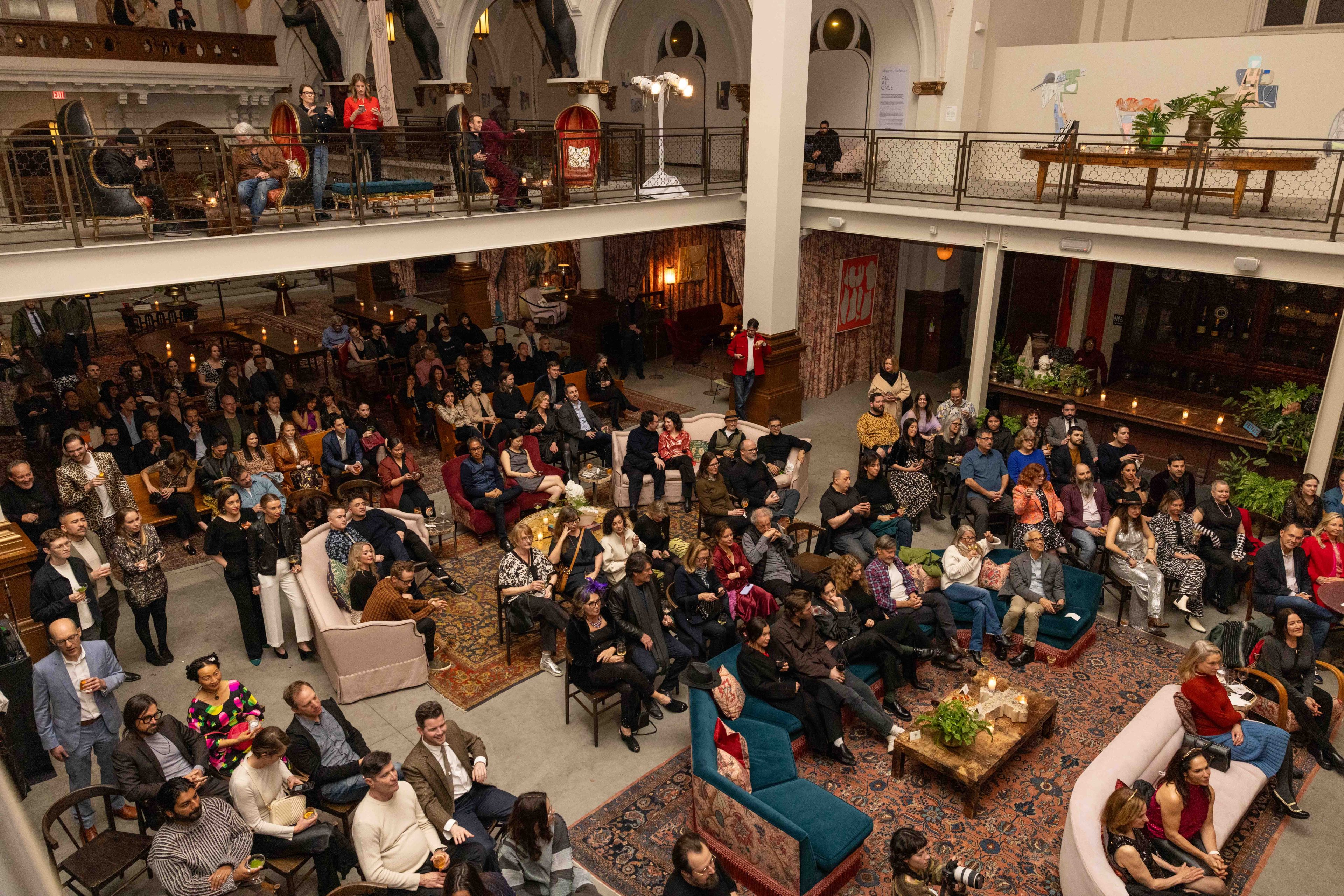 A large group of people is seated in a spacious, elegantly decorated room, attentively watching an unseen event, with some people on a balcony above.