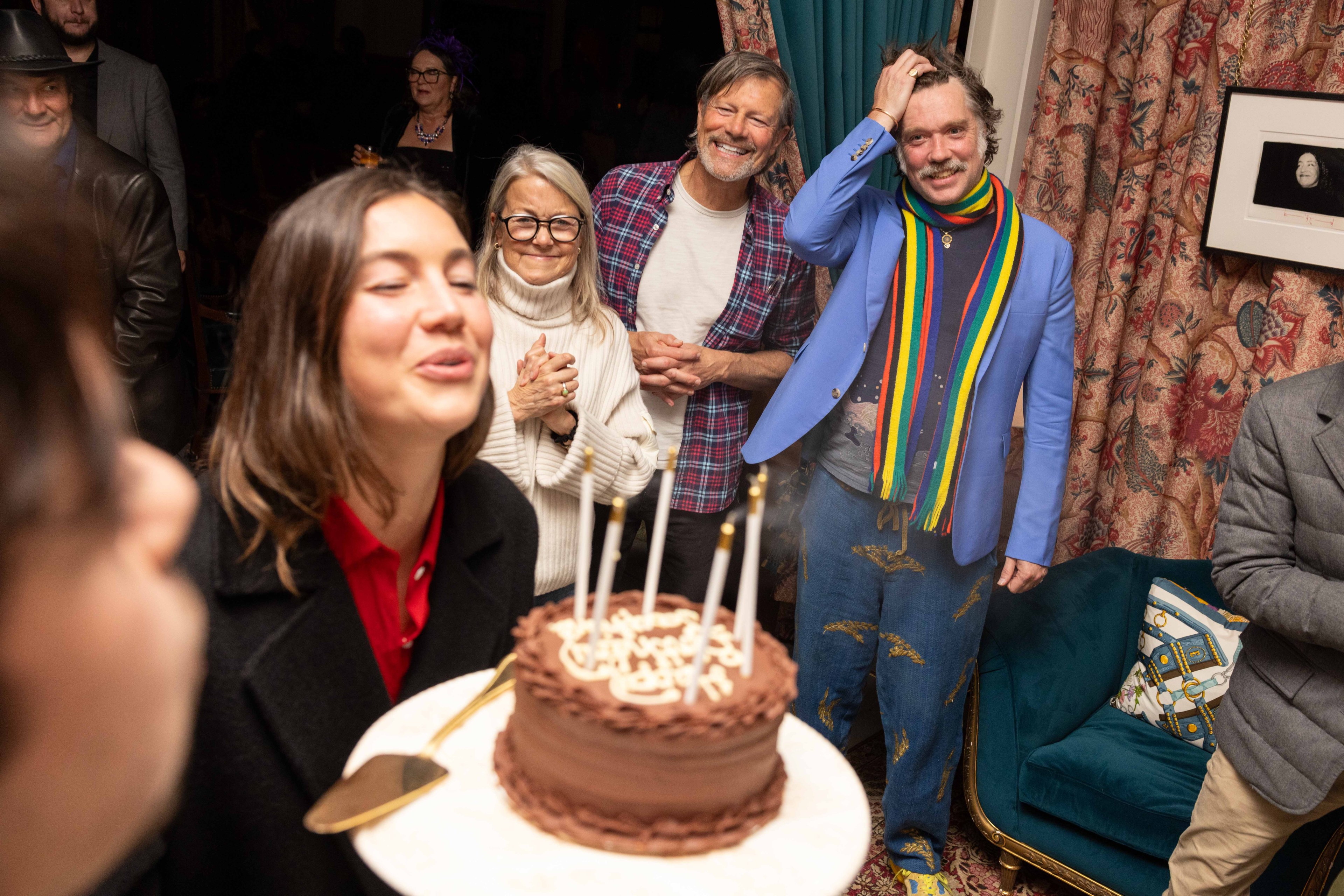 A group of people celebrates indoors. A woman holds a chocolate cake with candles. Others smile and watch, with festive attire and decorations around them.