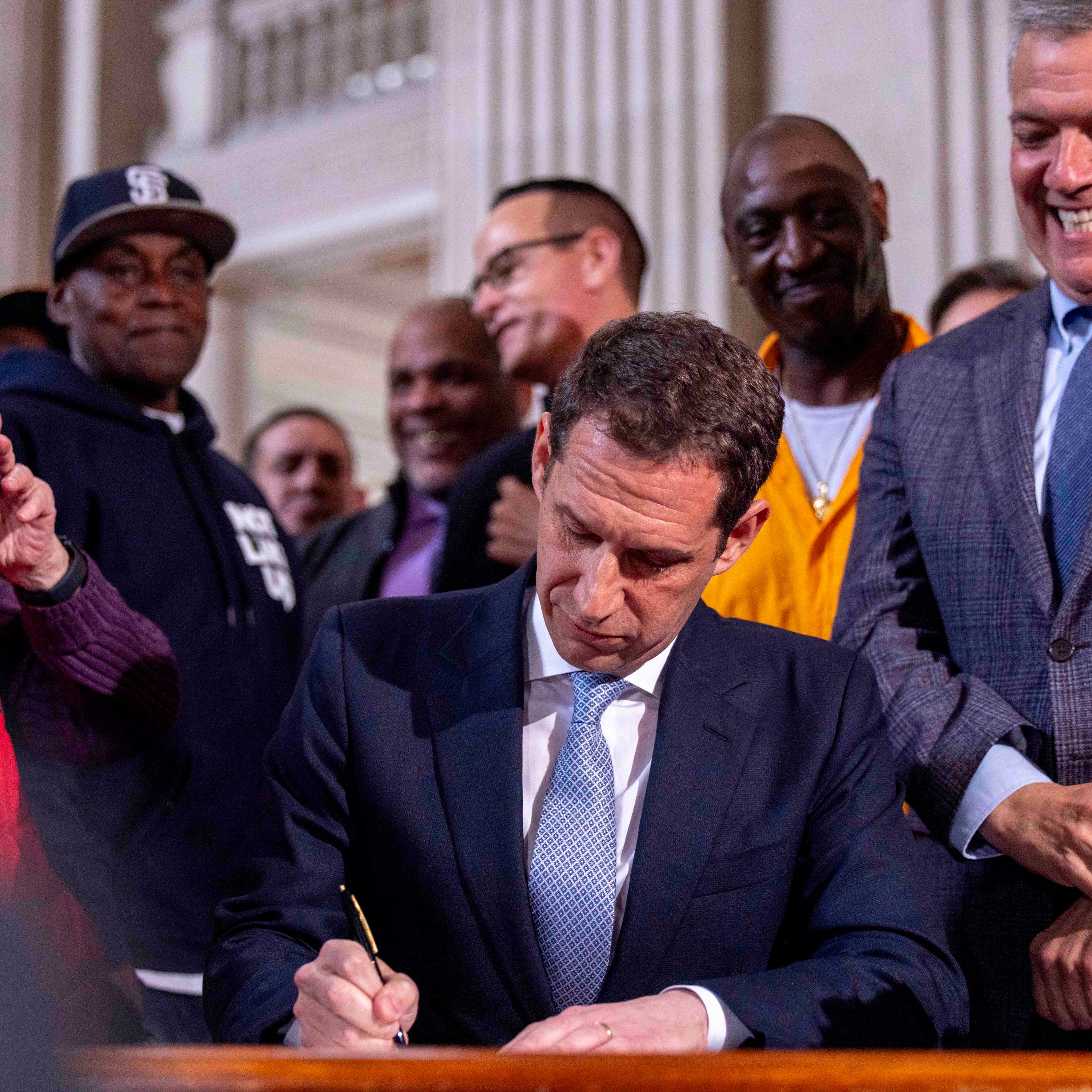A man in a suit is signing a document at a desk, surrounded by a smiling group of people, including someone taking a photo with a smartphone.