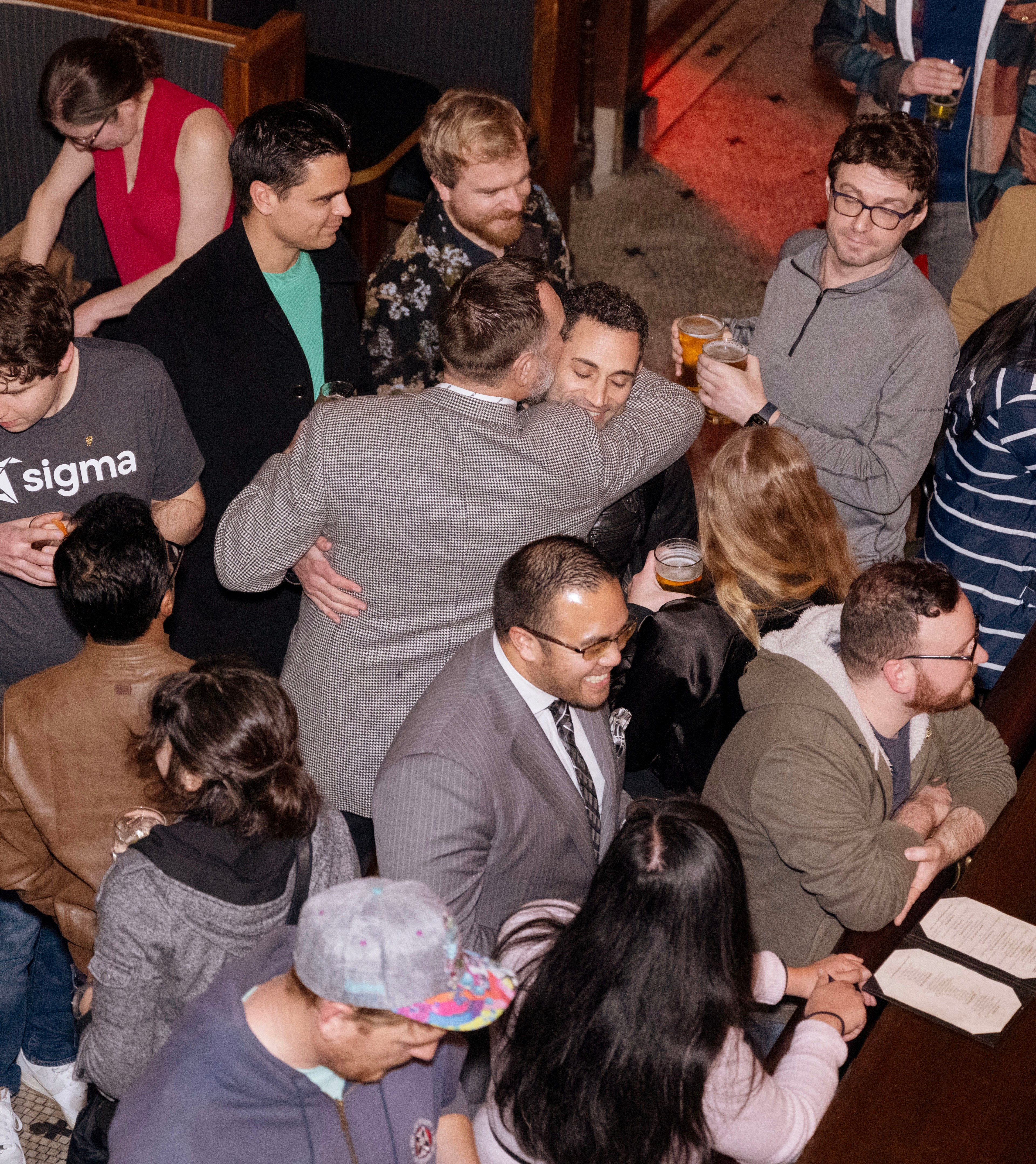 A crowded social gathering with people talking and holding drinks. Some are hugging or laughing, while others are looking at menus or engaged in conversations.