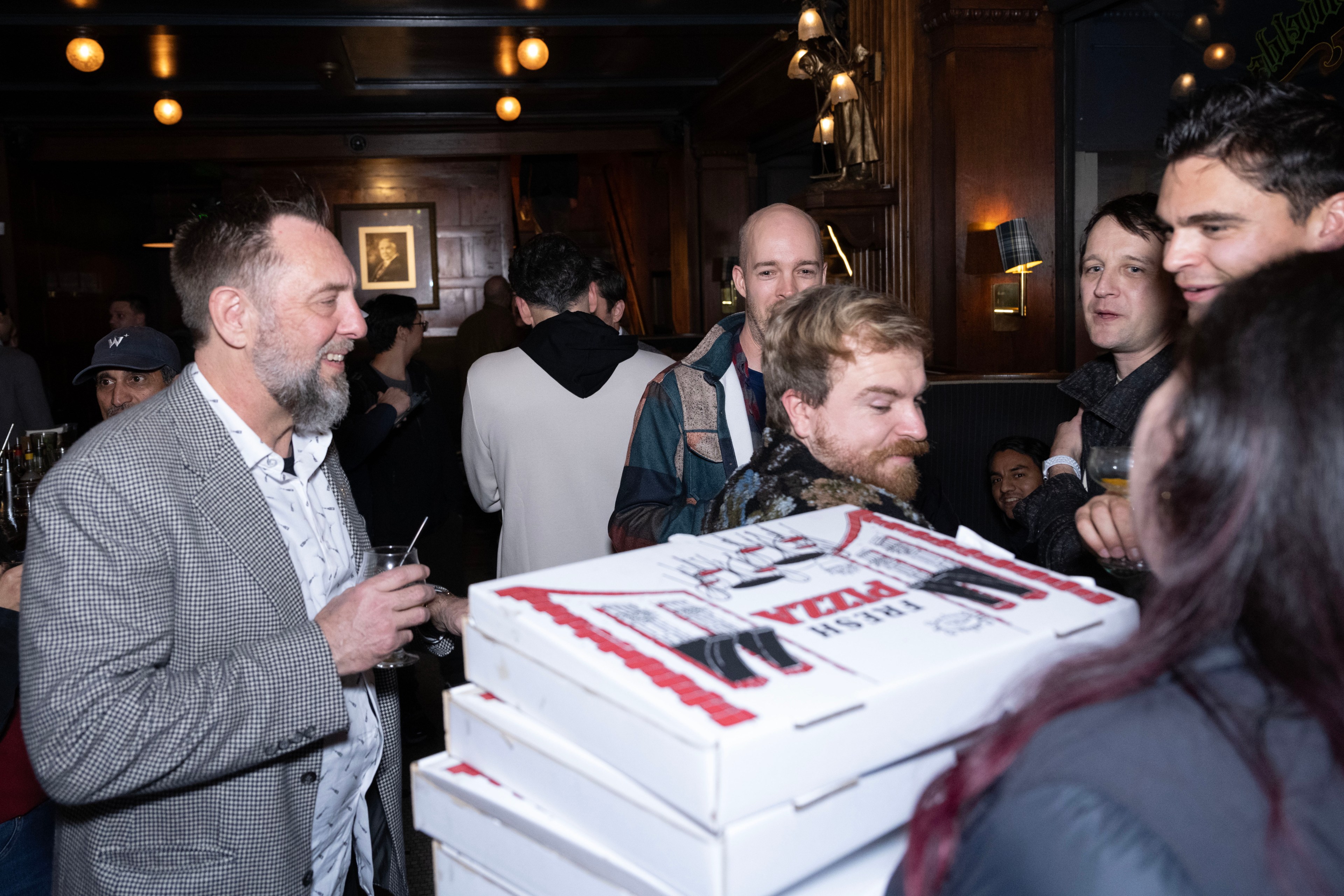 A group of people in a dimly lit room look at a pile of pizza boxes. A man in a checkered jacket and a woman with pink-tipped hair are prominent in the scene.