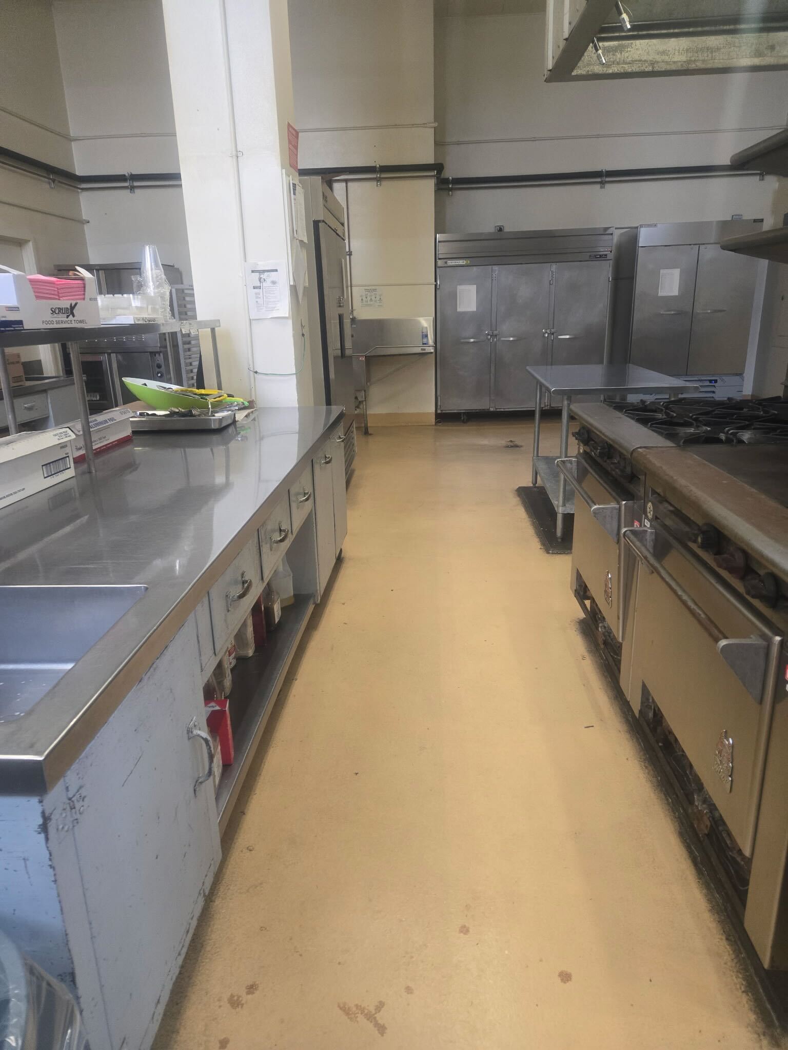 A commercial kitchen with stainless steel countertops, sinks, cooking equipment, and large silver refrigerators. The floor is clean and beige.