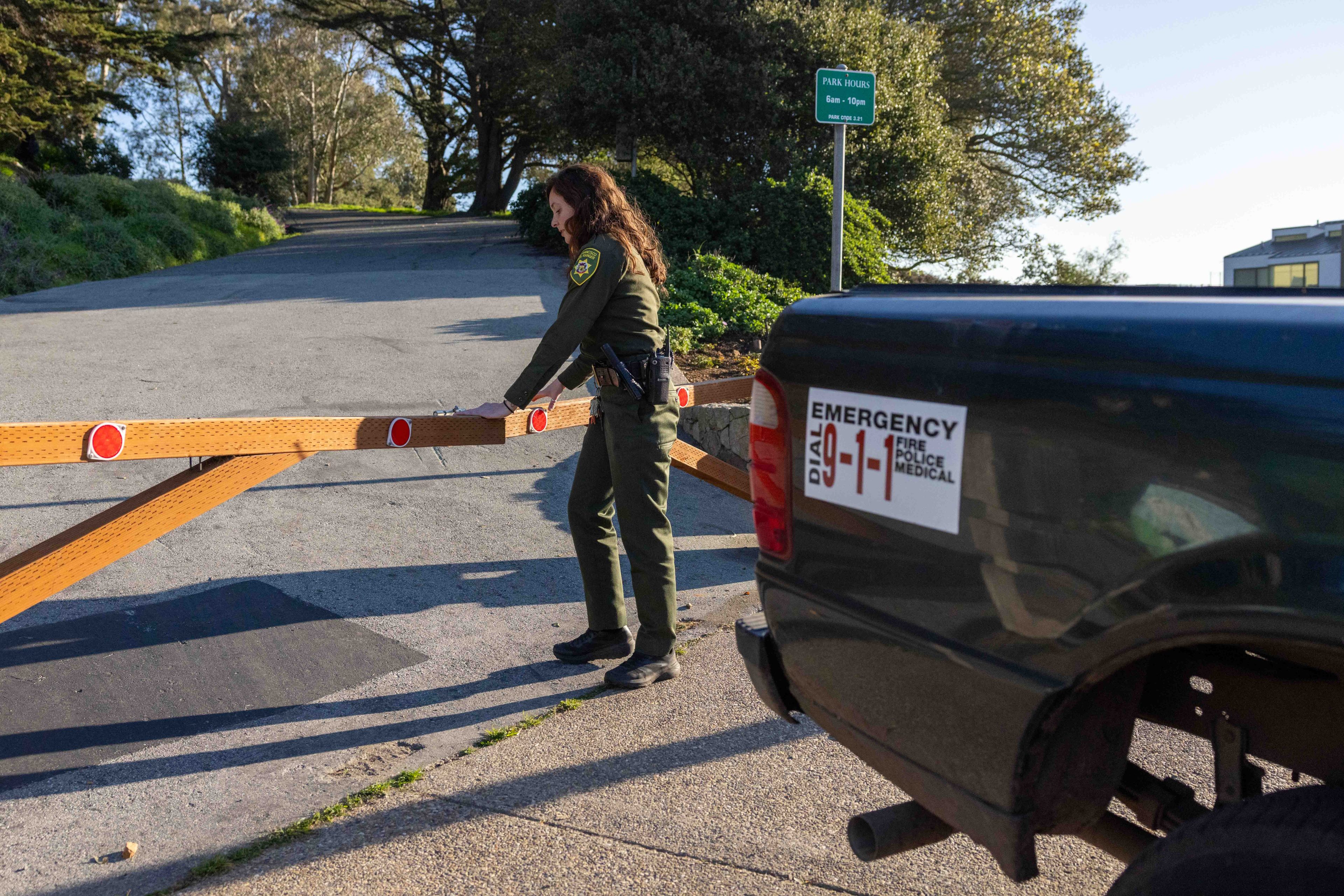 A park ranger in uniform is closing a wooden gate. A sign on a nearby vehicle reads &quot;EMERGENCY DIAL 9-1-1&quot;. Trees and a sign with park hours are visible.