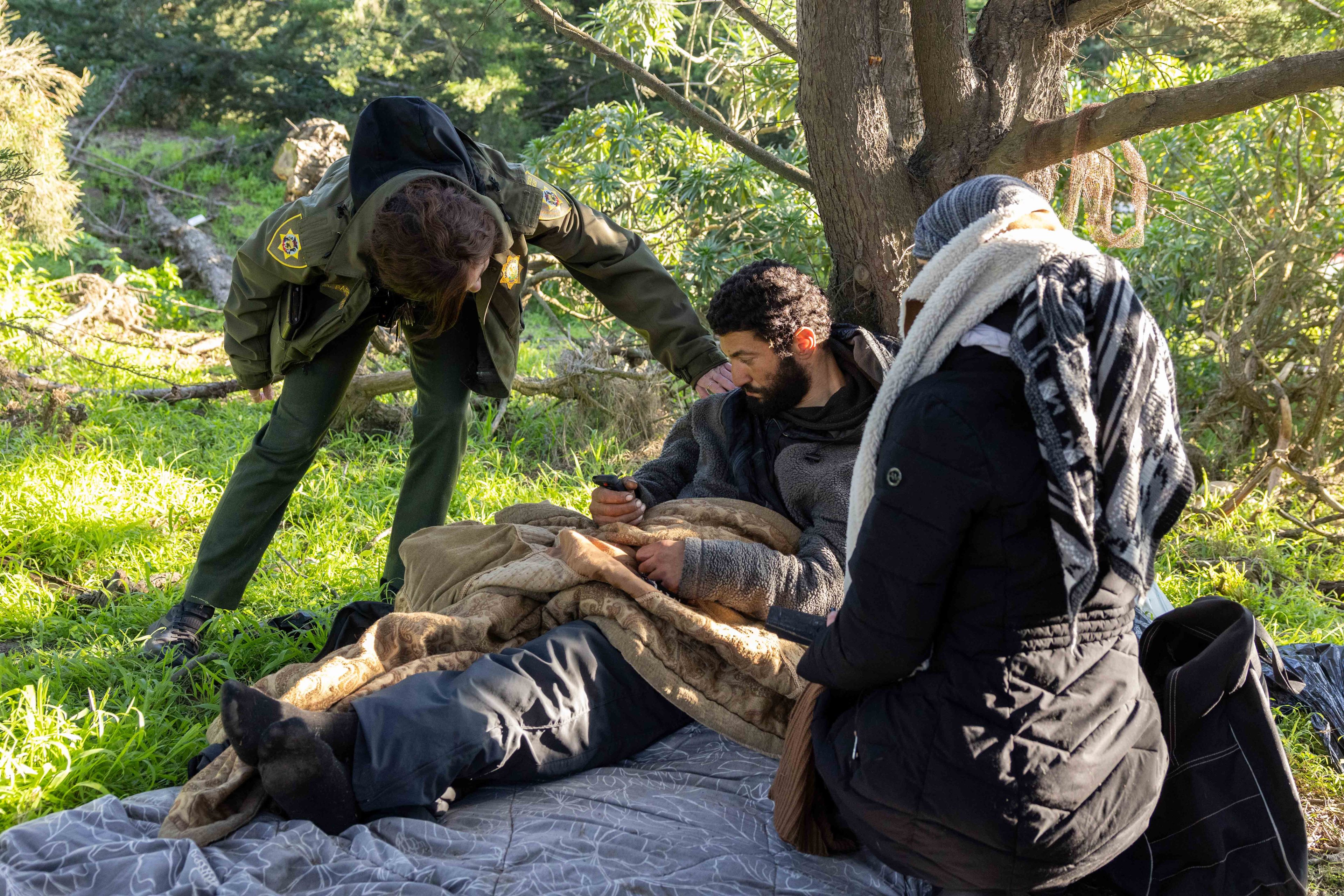 A person in uniform checks on two individuals wrapped in blankets on the ground in a forest. Bright sunlight filters through the trees surrounding them.