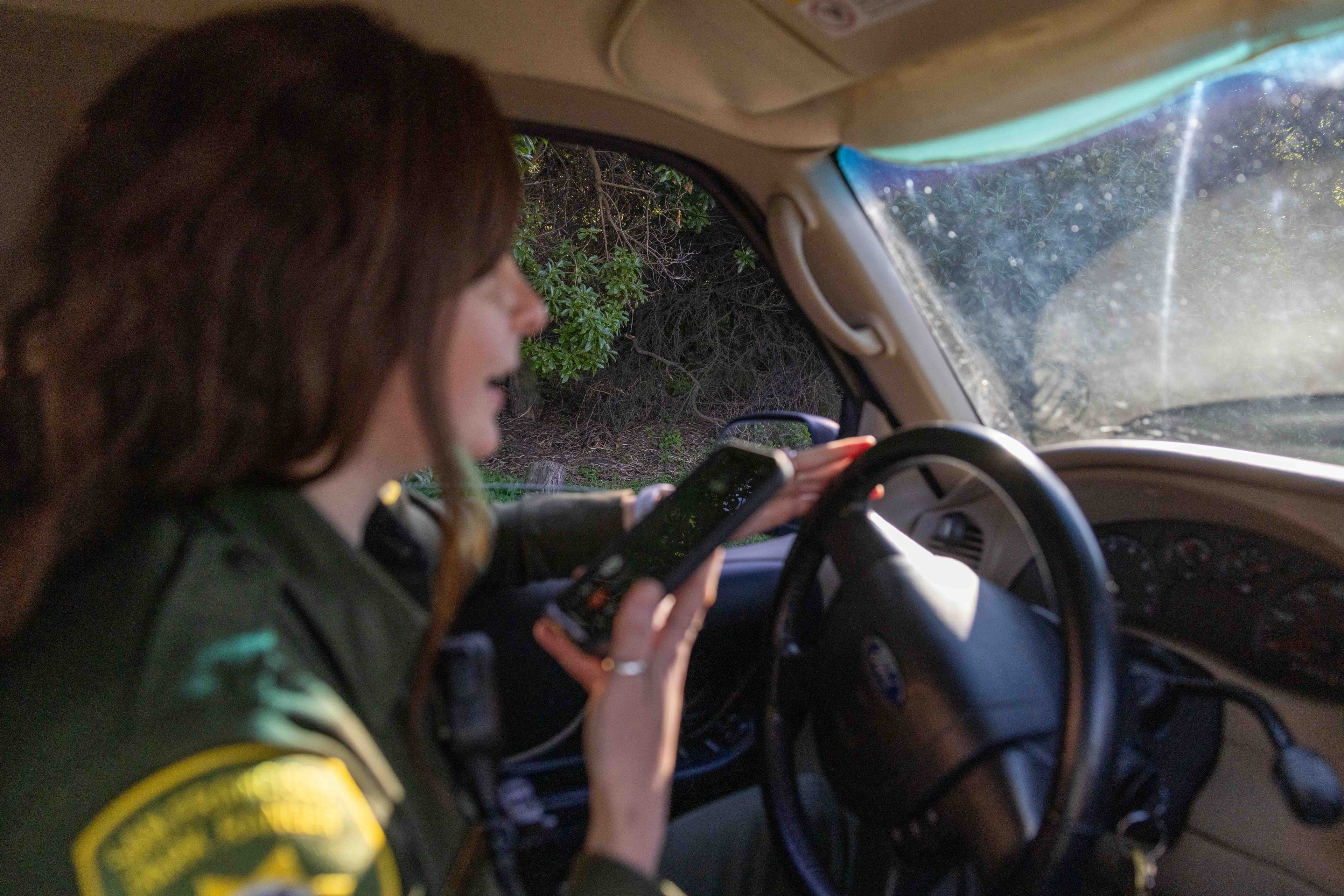 A person in a uniform is seated in a car, holding a smartphone and speaking. Sunlight illuminates the interior, and foliage is visible through the window.