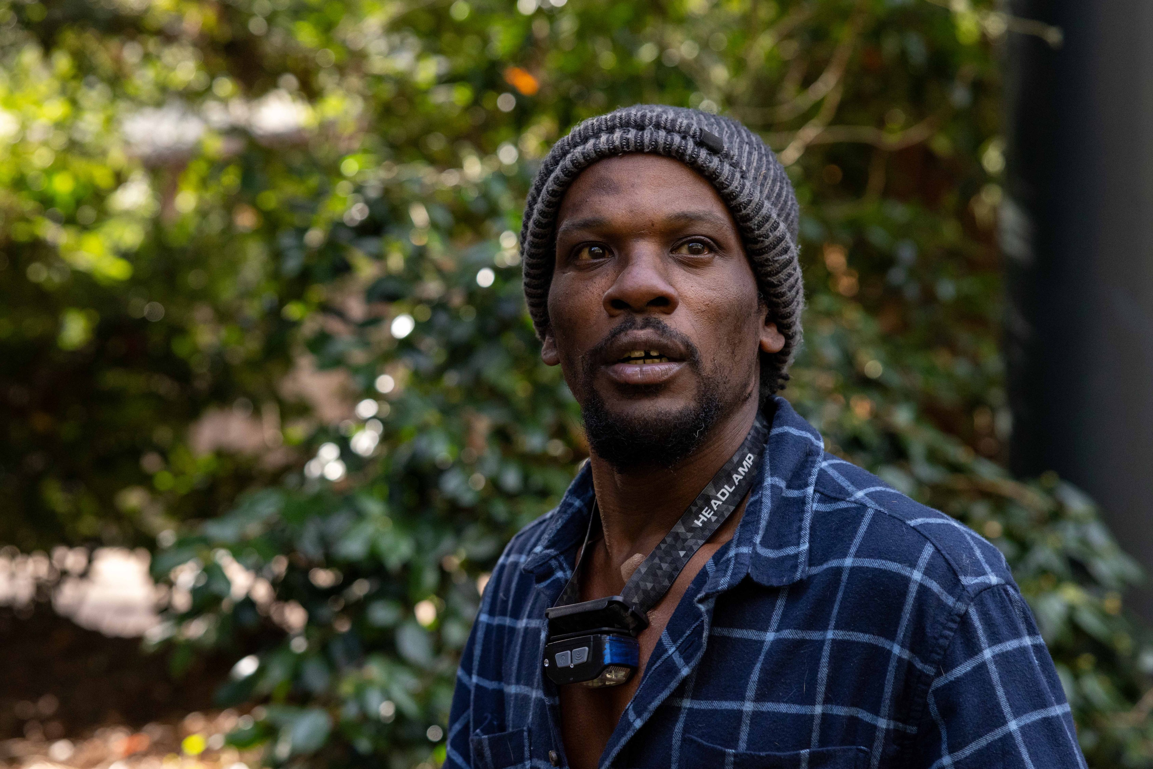 A man in a gray knitted hat and blue plaid shirt stands outdoors, wearing a headlamp around his neck. The background is blurred green foliage.