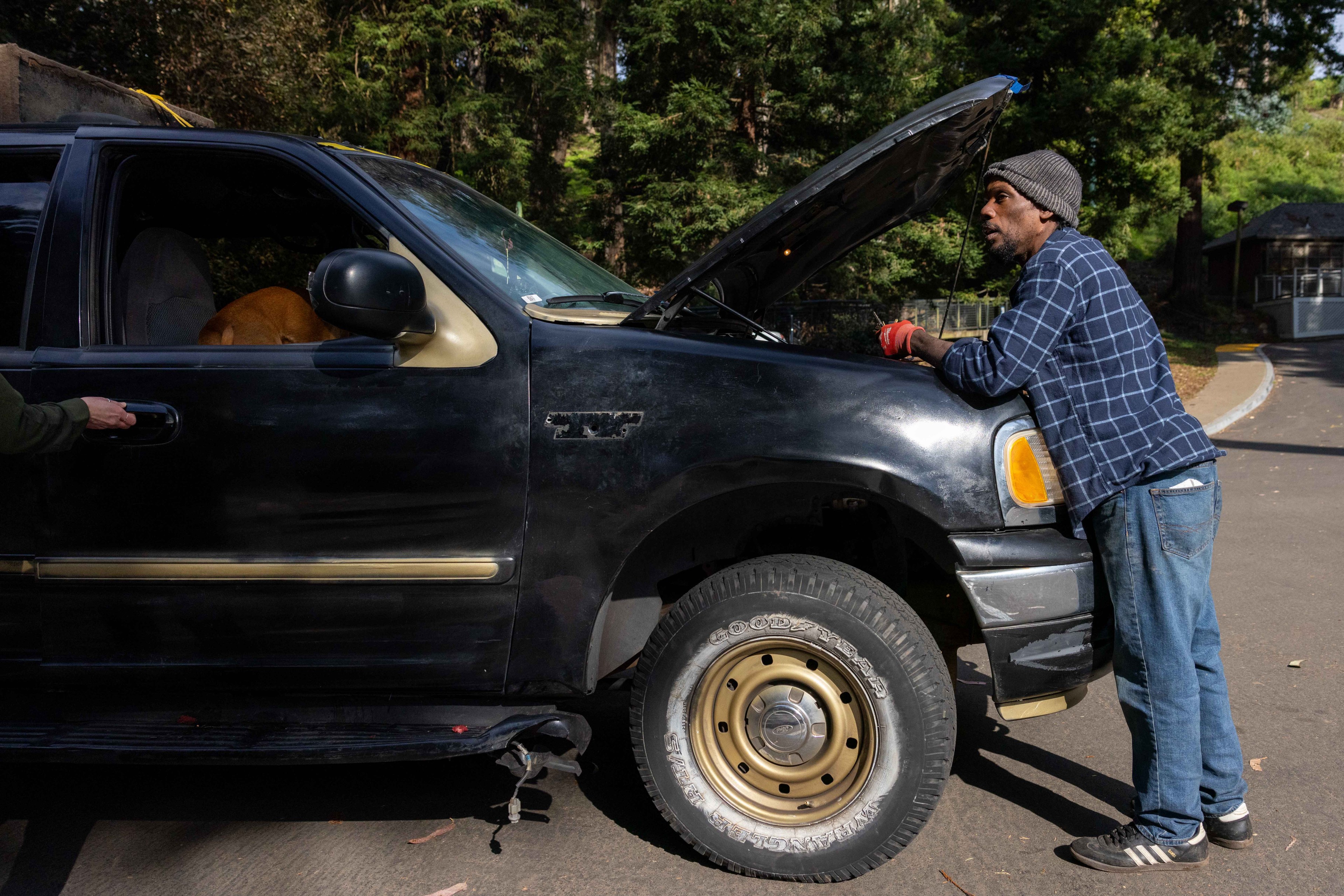 A man in a plaid shirt and beanie leans over the open hood of a black SUV. A dog is visible inside the car, and another person is opening the door.