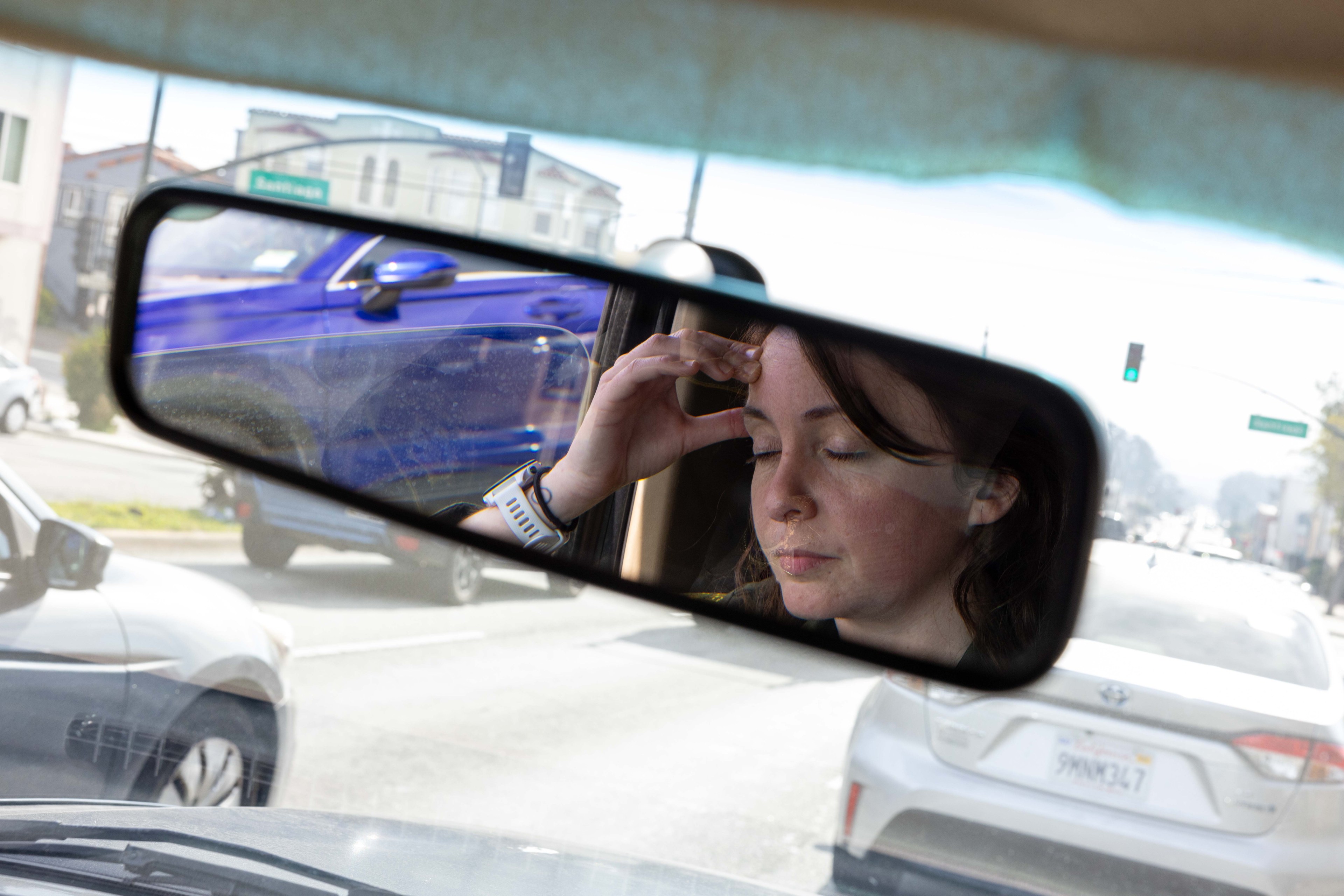 A woman in a rearview mirror holds her forehead, eyes closed, with cars visible through the window. She appears thoughtful as traffic moves around.