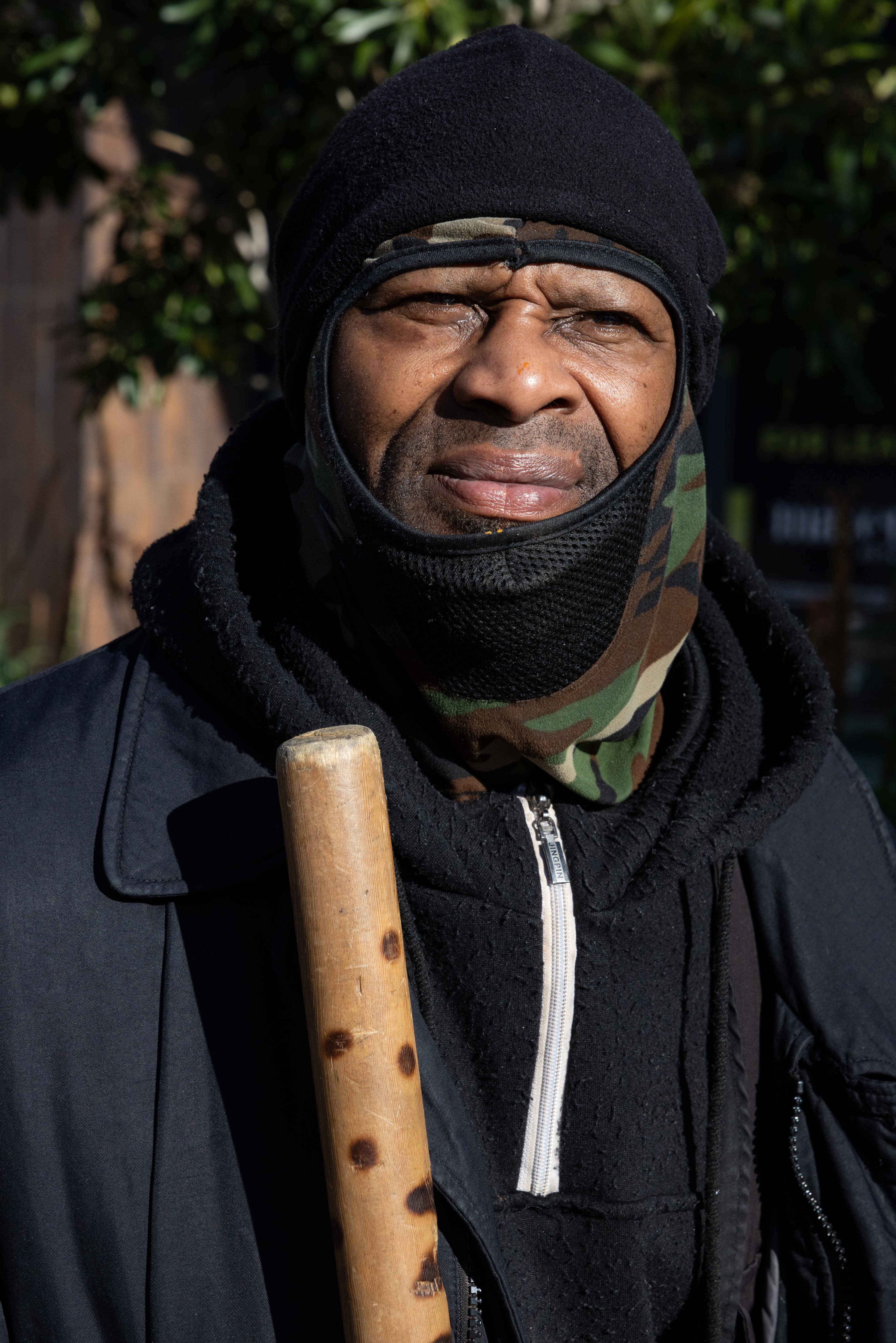 A person bundled in a black hood and camo mask squints in sunlight, holding a wooden stick. They wear a black jacket and a zip-up sweater.