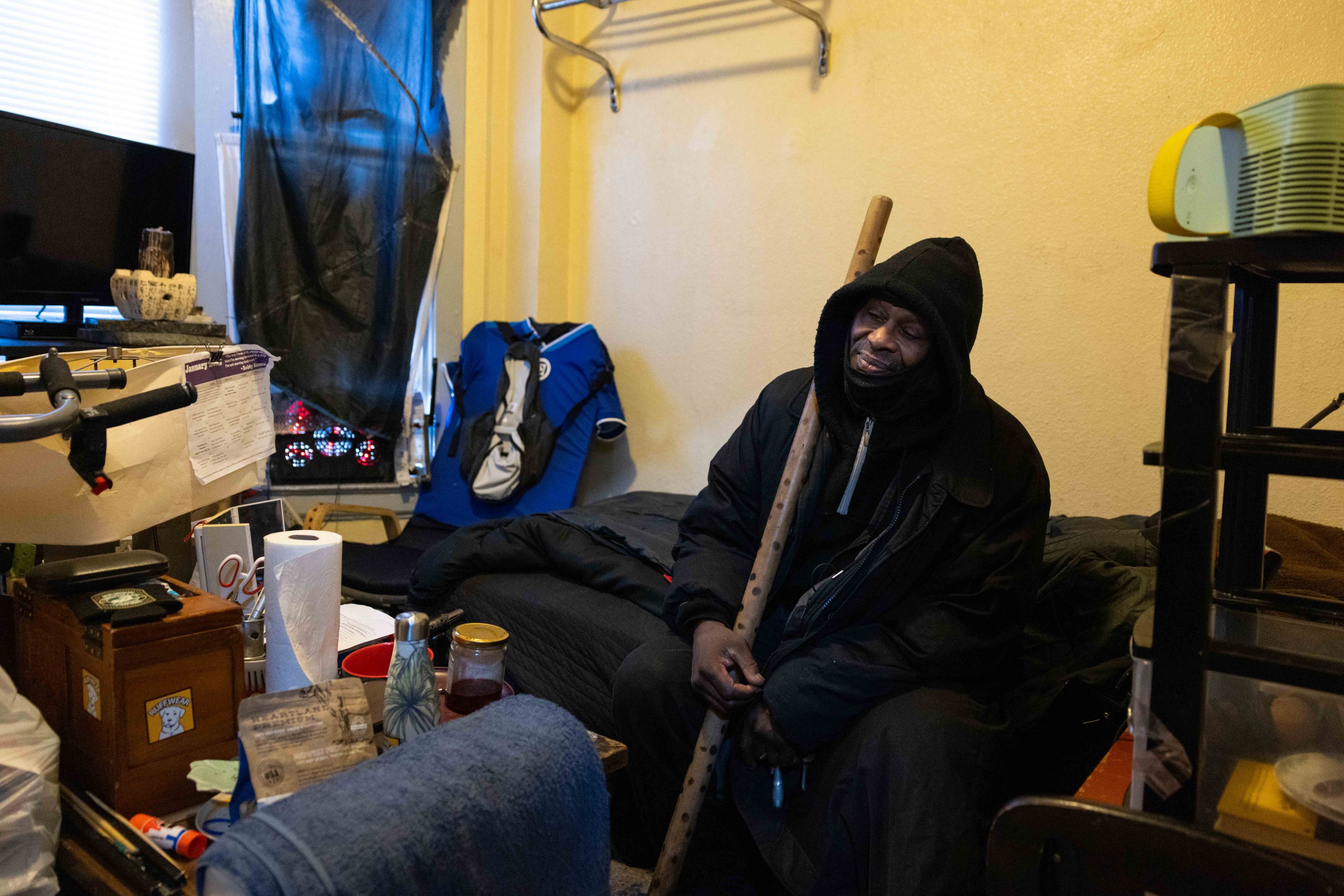 A person wearing a hooded coat sits on a small bed in a cluttered room, holding a wooden stick. Various items are scattered, and a small TV is visible.