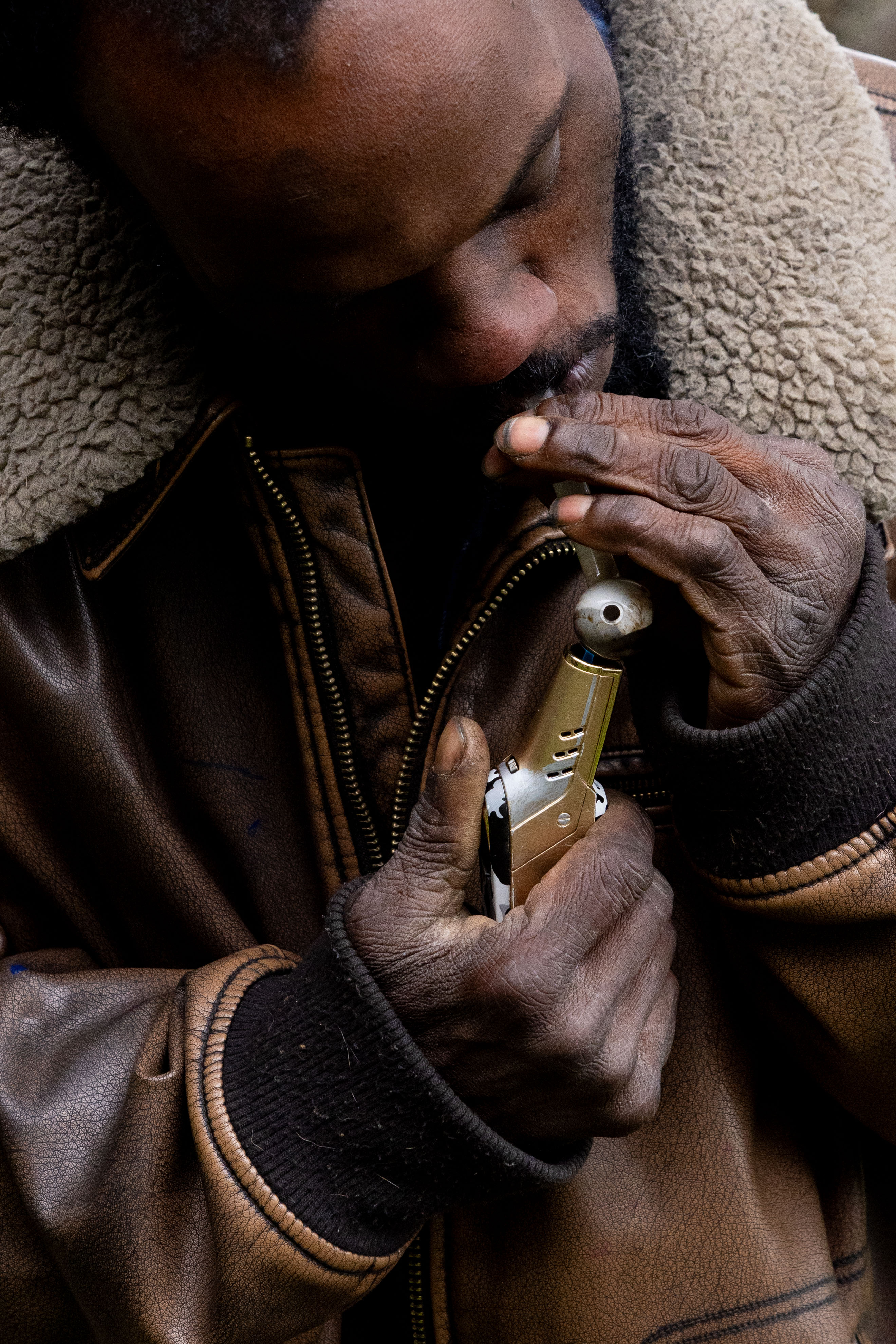 A person wearing a brown jacket with a shearling collar is holding a pipe and lighter, appearing to light the pipe with the flame.