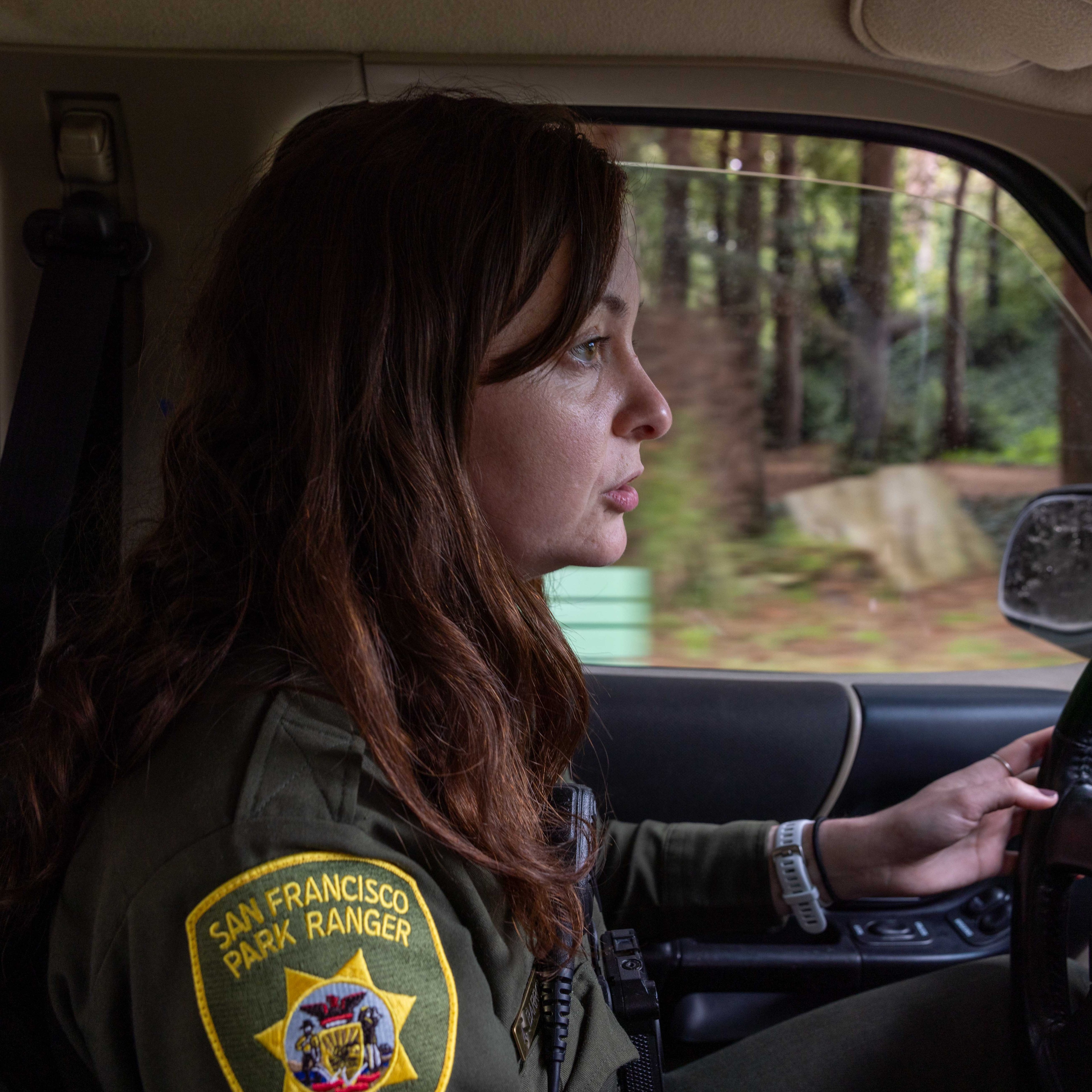 A person wearing a San Francisco Park Ranger uniform is driving a vehicle. The scene outside the window shows trees and greenery, suggesting a natural setting.
