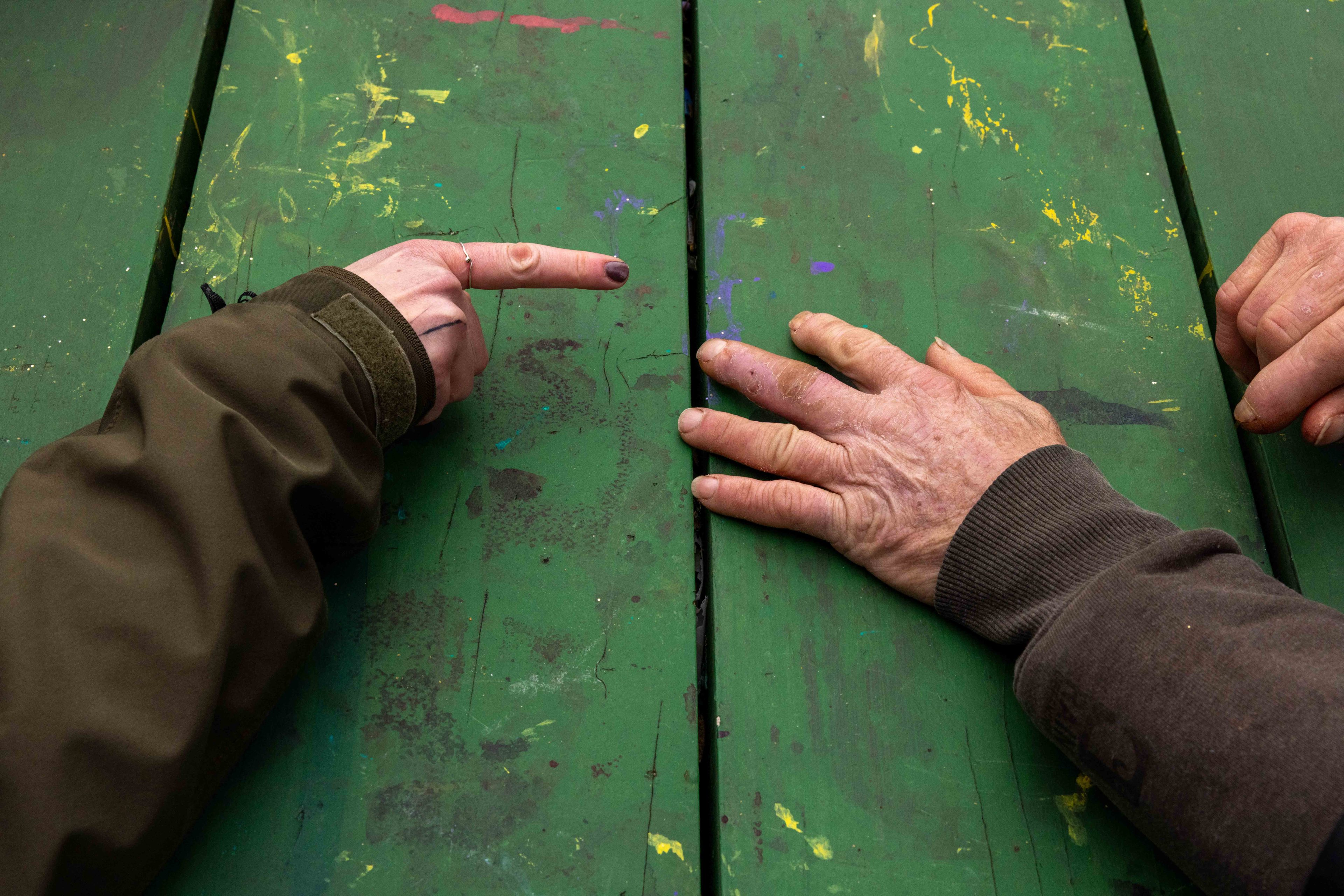 Two hands rest on a weathered, paint-spattered green table. One hand points, while the other is flat, showing different textures and colors.