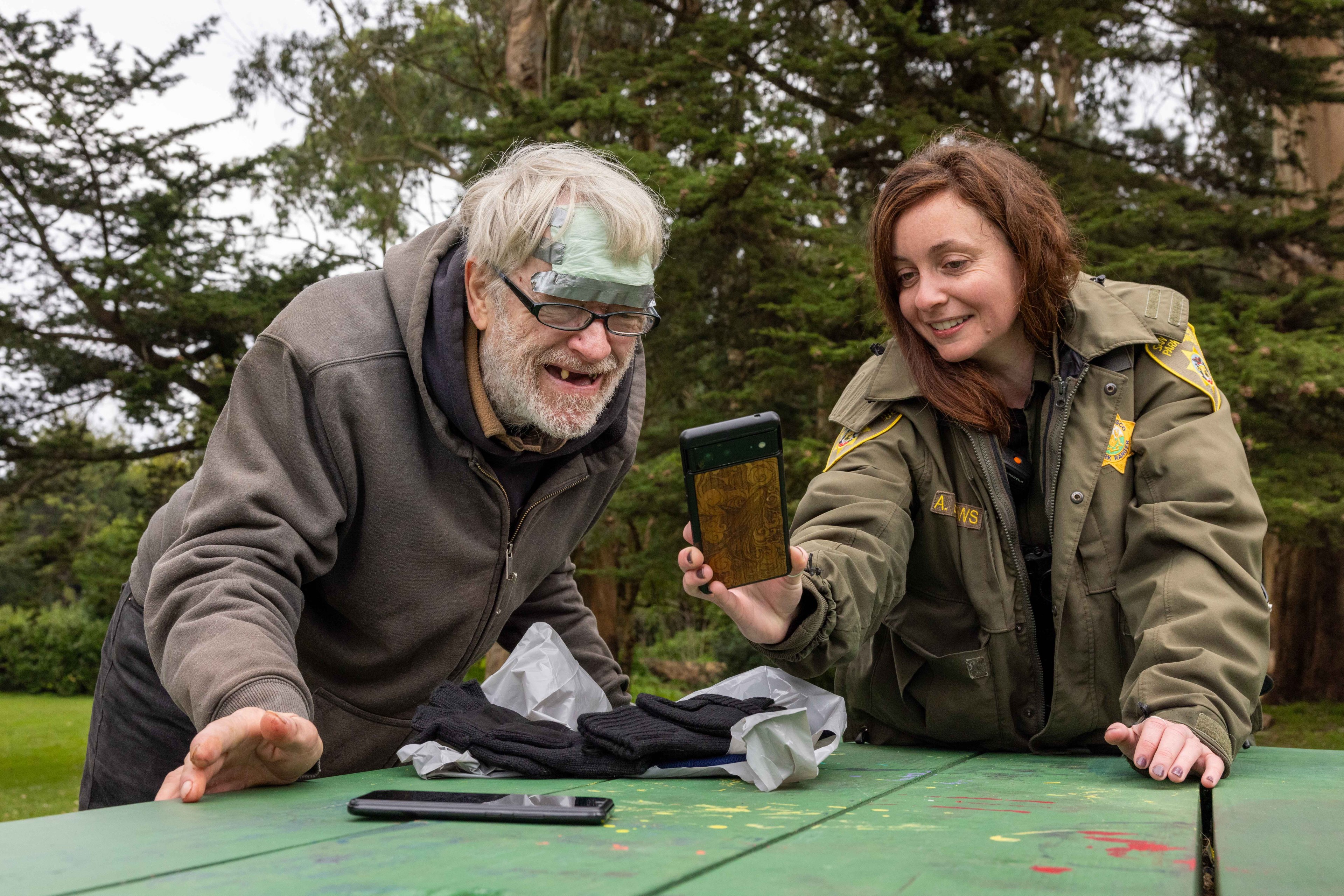 A smiling man in a hoodie and a woman in a uniform share a light moment outdoors, looking at a phone with a wooden-patterned case on a picnic table.