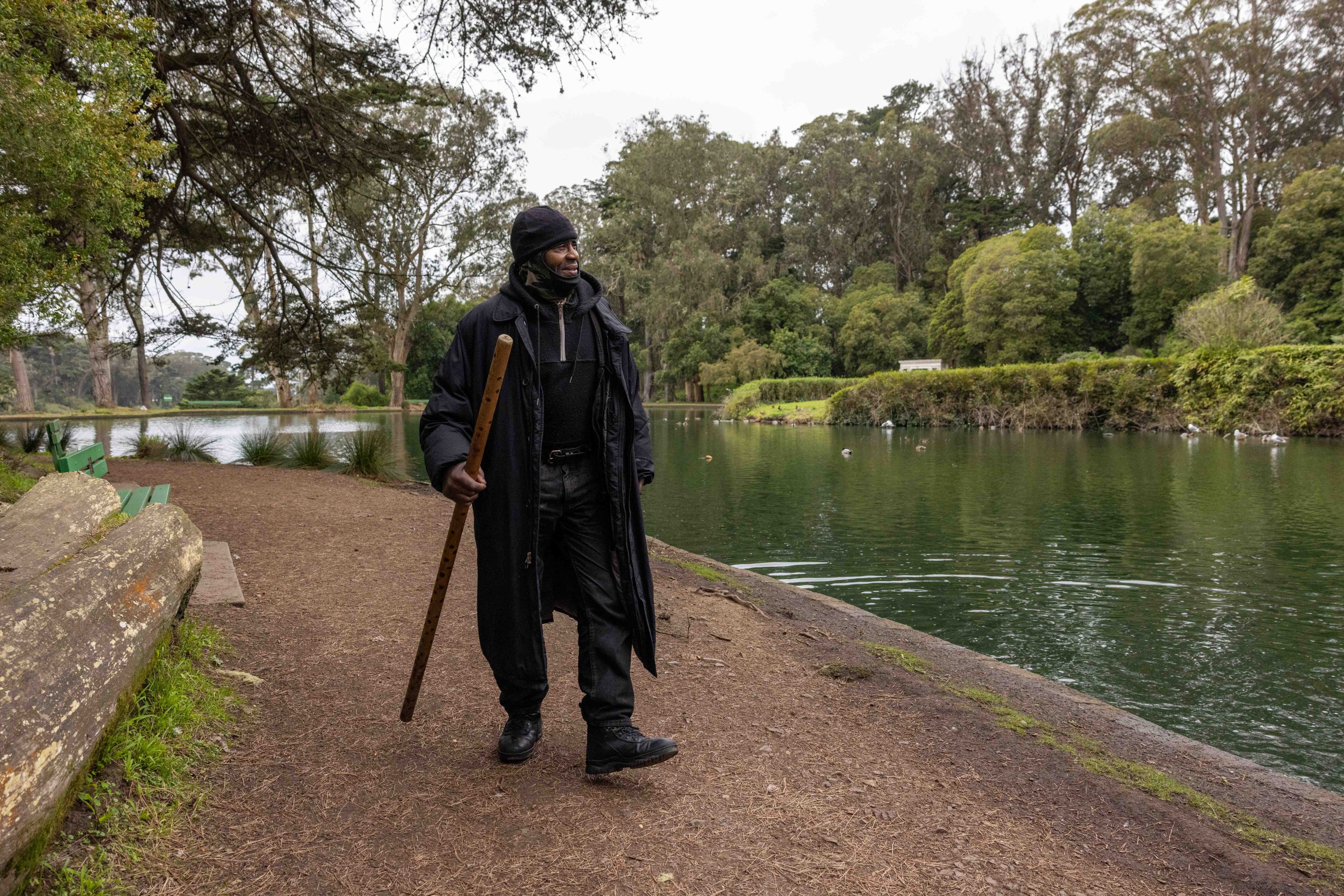 A person dressed in dark clothing and a beanie walks with a wooden staff beside a calm pond, surrounded by trees and greenery.