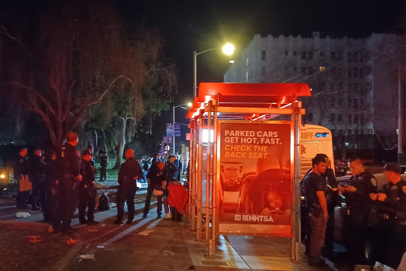 A nighttime scene with a group of police officers standing near a brightly lit bus stop. The bus stop displays an ad about parked cars getting hot quickly.