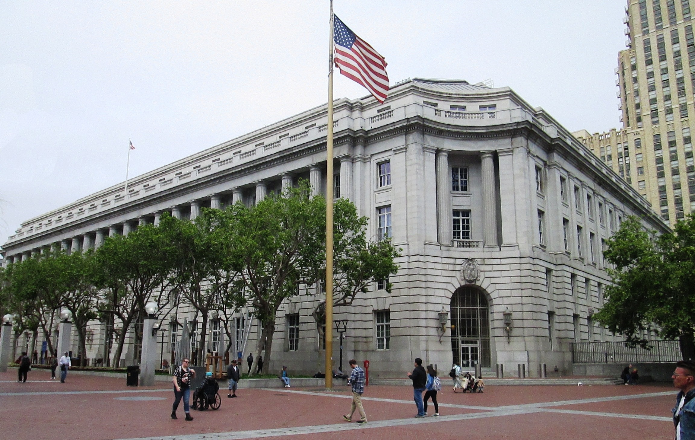 A large neoclassical building with columns is in the background. An American flag flies prominently in front. People walk and gather in the open plaza.