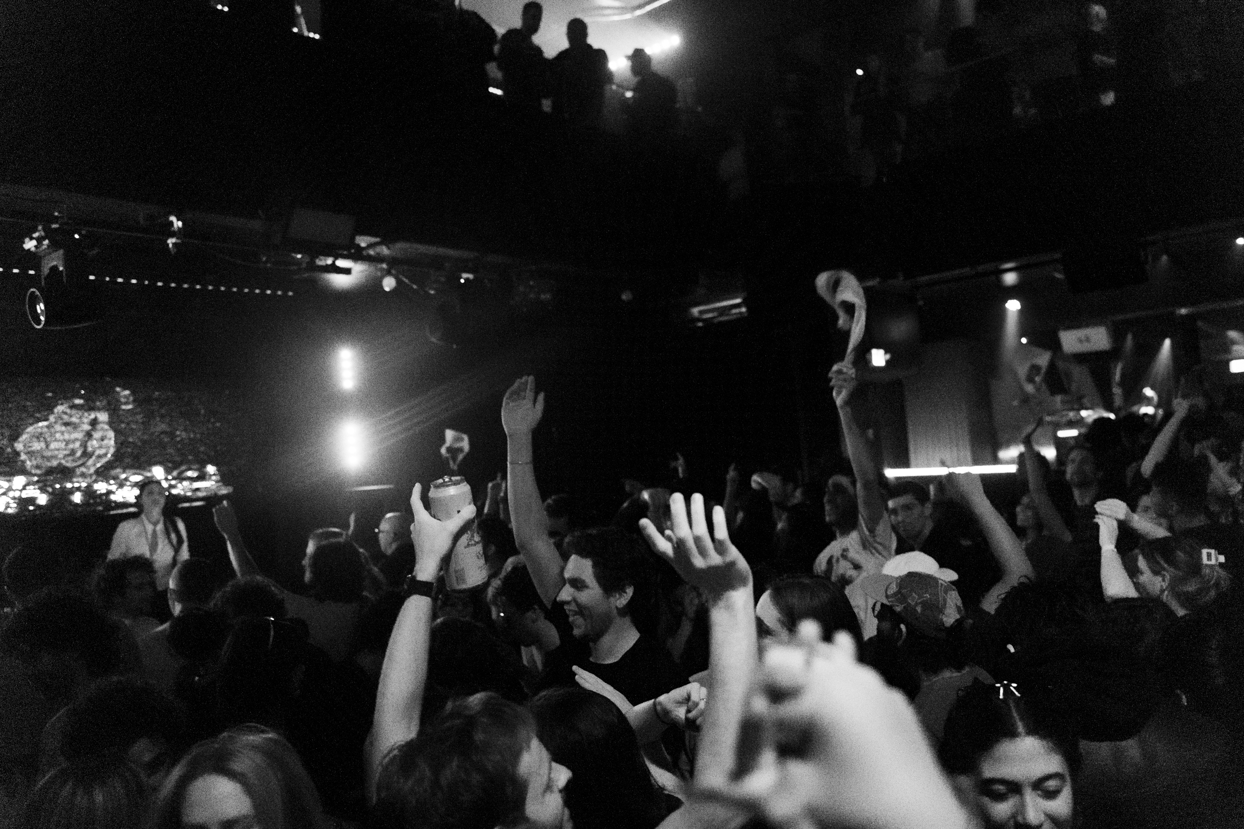 A lively, crowded club scene with people raising their hands, some holding drinks, and the atmosphere is dimly lit with lights and shadows enhancing the energy.