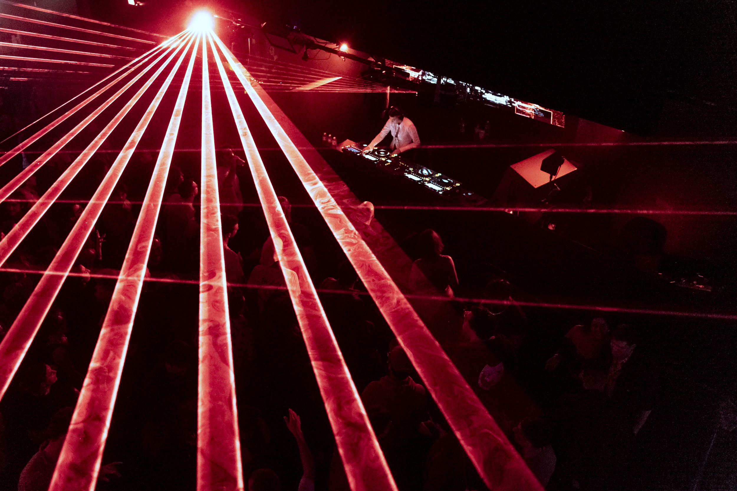 A DJ performs on stage with red lasers shining above a dancing crowd, creating a vibrant, energetic atmosphere in a dimly lit venue.