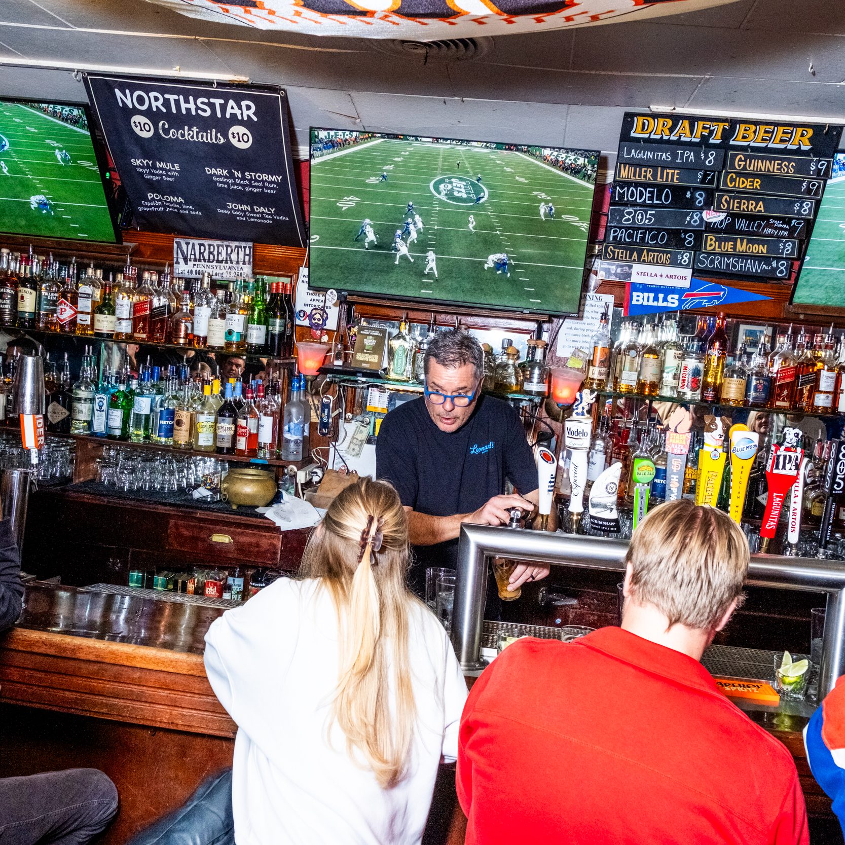 A bartender is serving drinks in a lively bar with sports on TV screens above. The bar is stocked with various bottles and draft beer options listed.