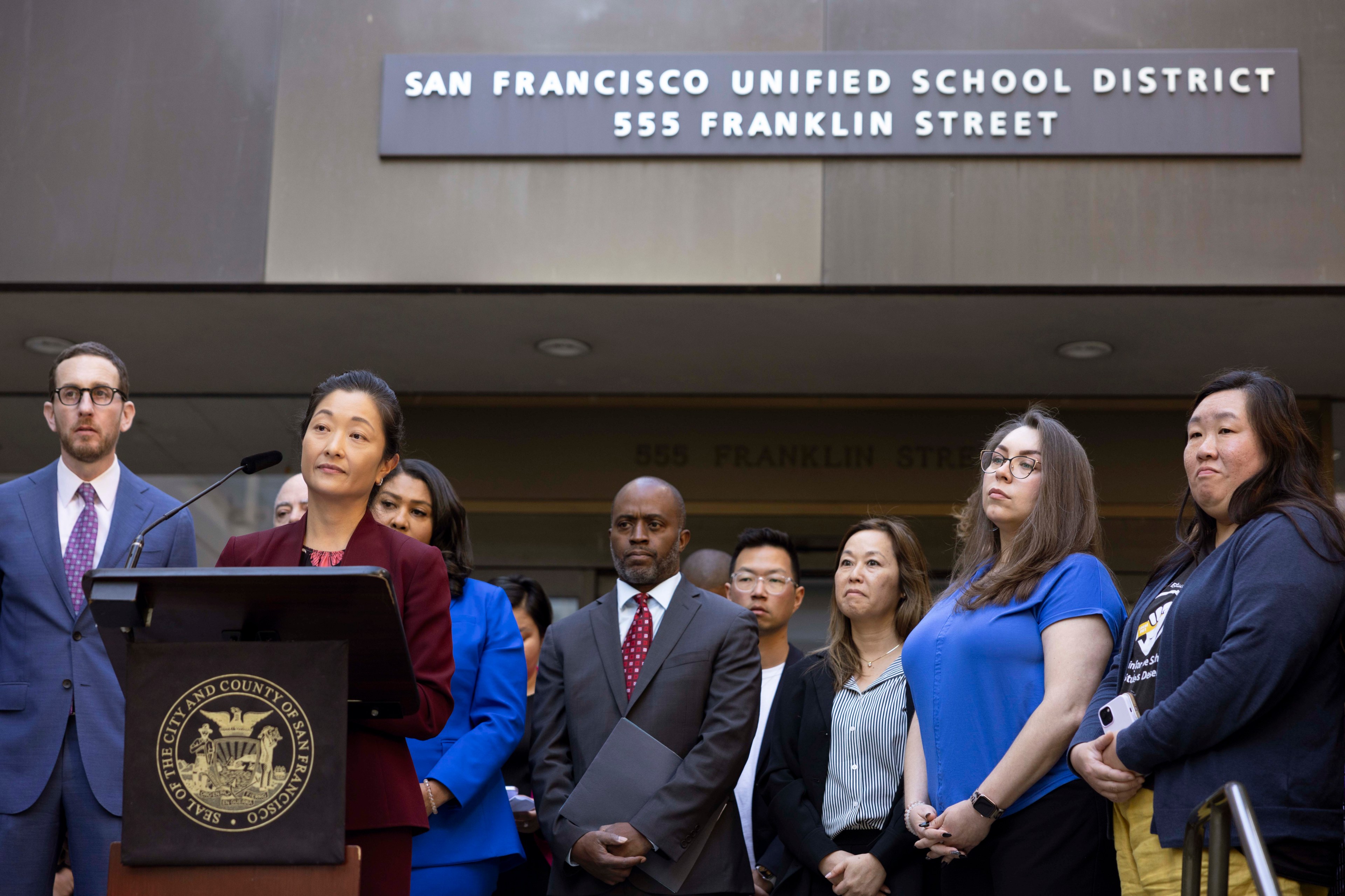 A group of individuals stands outside the San Francisco Unified School District building. A woman at a podium is speaking, surrounded by attentive people.