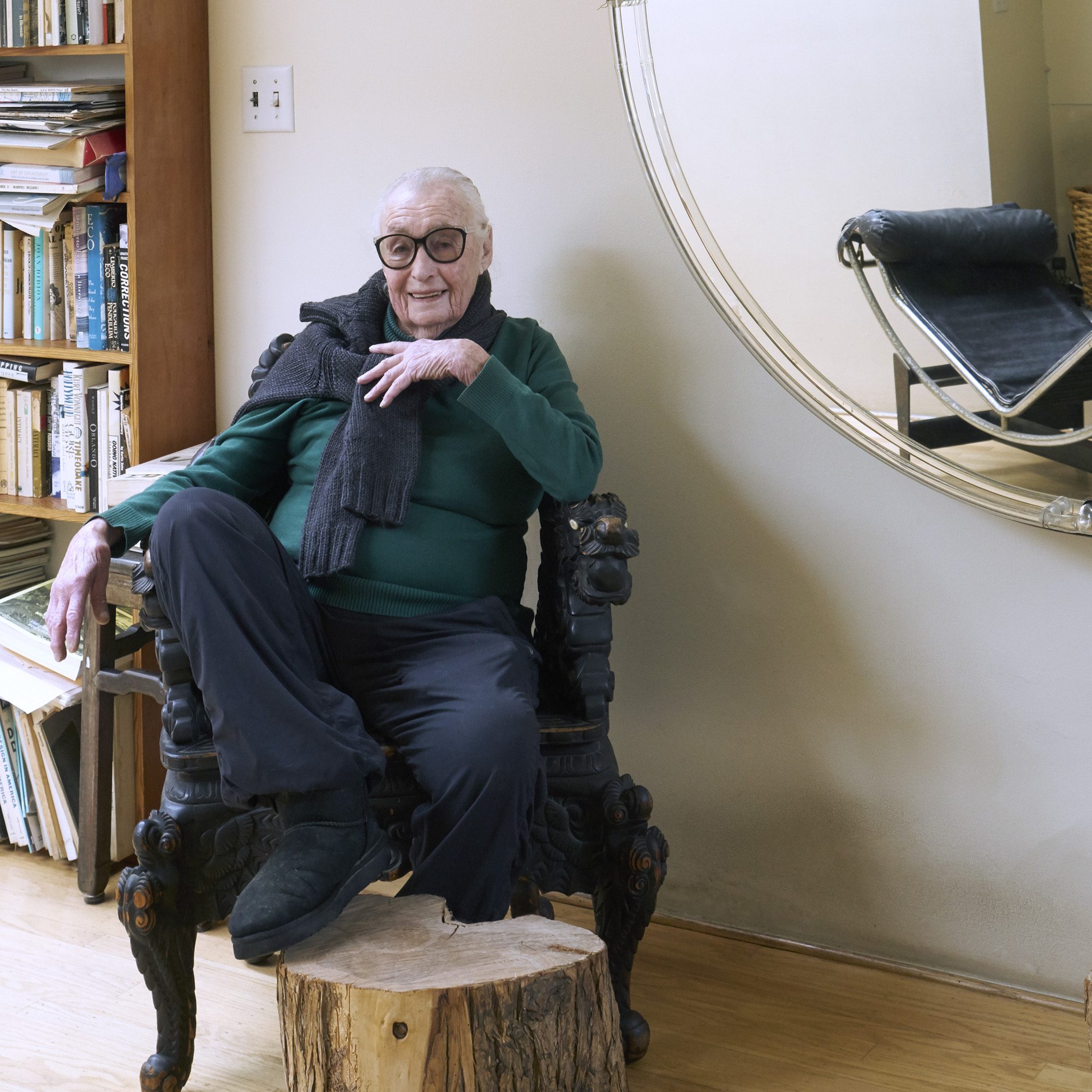 An elderly person with glasses is seated in an ornate chair, wearing a green sweater and scarf. A large mirror and bookshelf with various books are in the background.