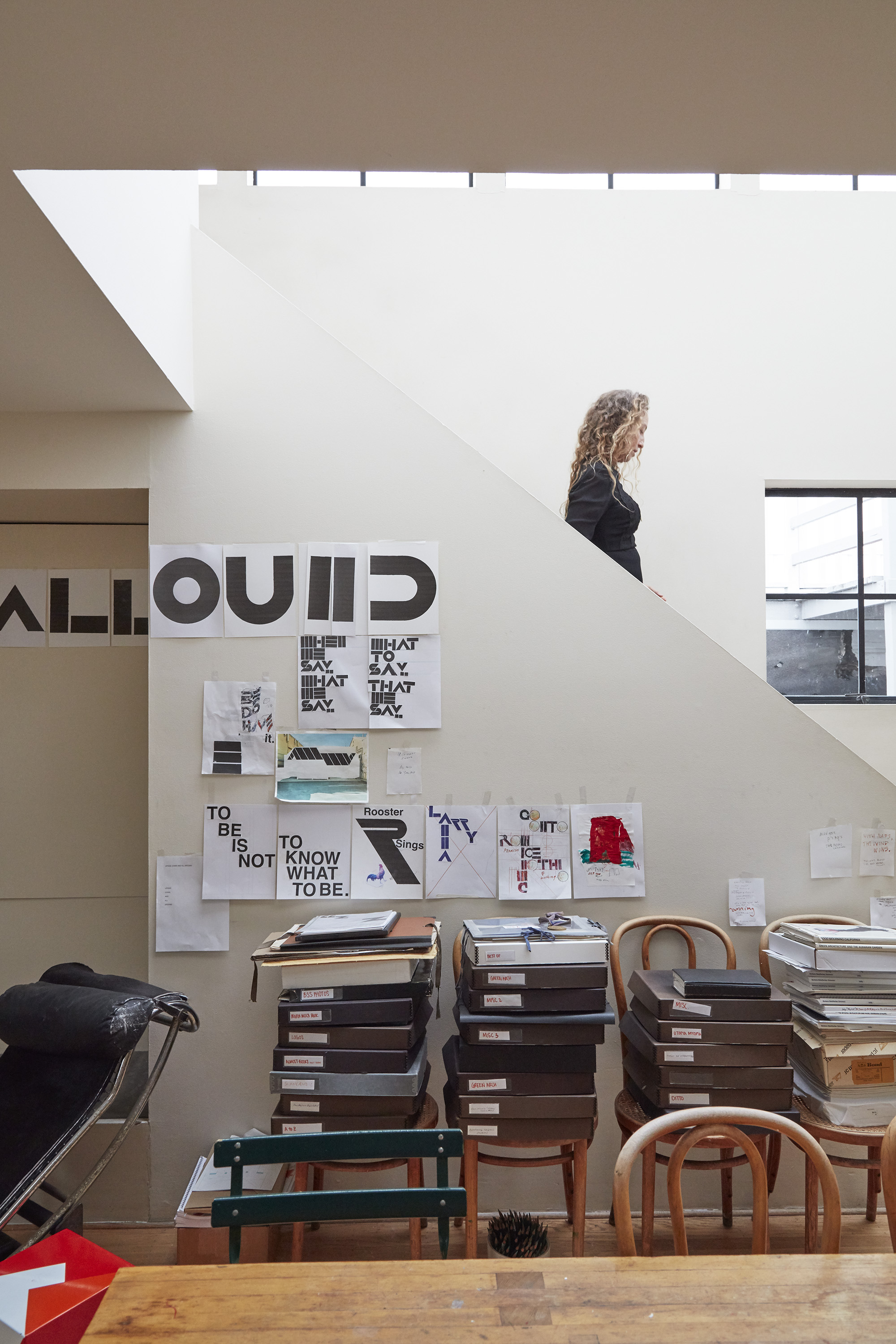 A woman with curly hair walks up a modern staircase in a well-lit room with art and papers on the wall. Stacks of boxes and chairs are below the art.