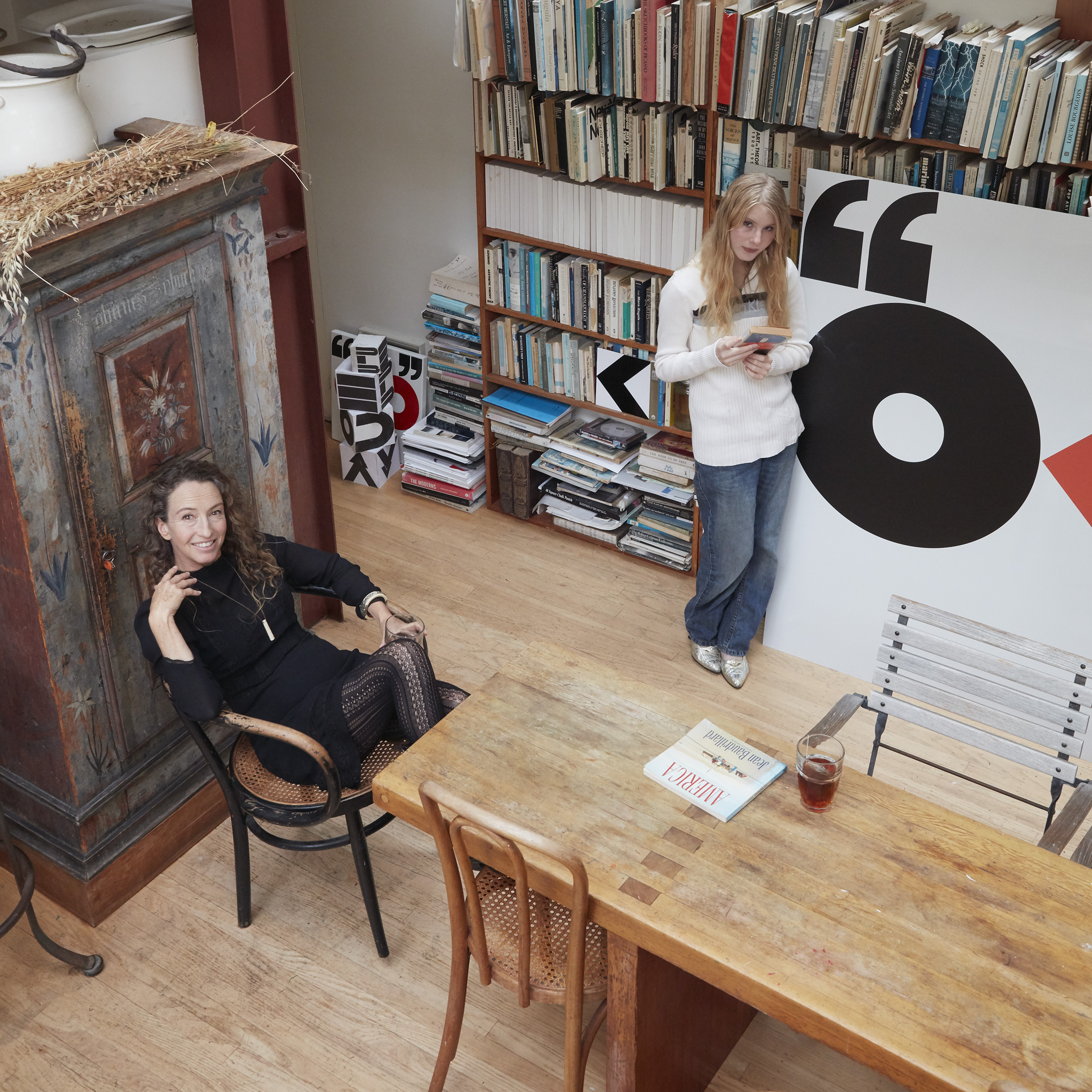 Two people are in a library, one sitting on a chair and the other leaning on a large art piece. Books fill shelves; a wooden table holds a magazine and drink.