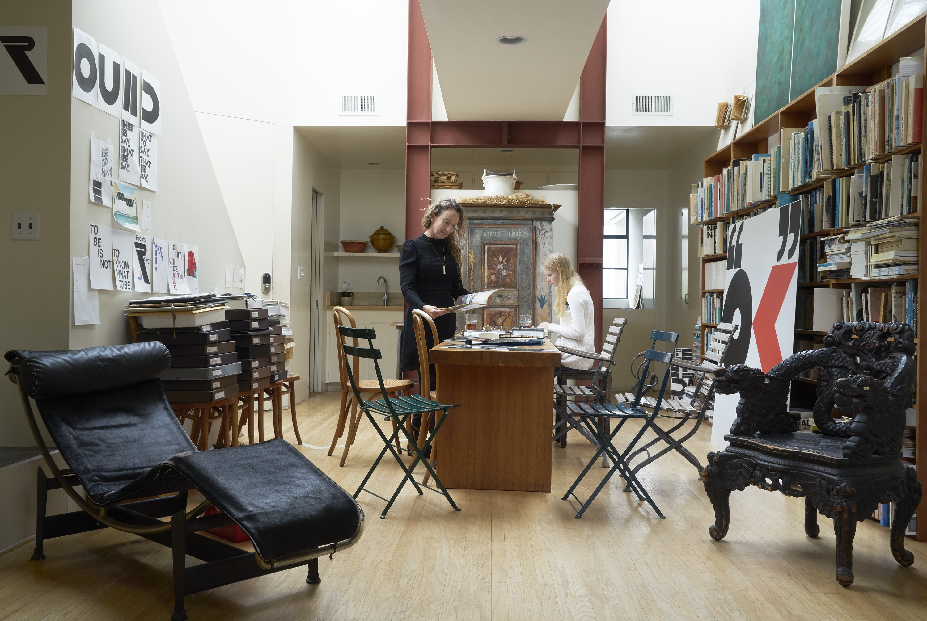 A spacious, well-lit room with two people working at a wooden table, surrounded by bookshelves, artwork on the walls, and various chairs, including a lounge chair.