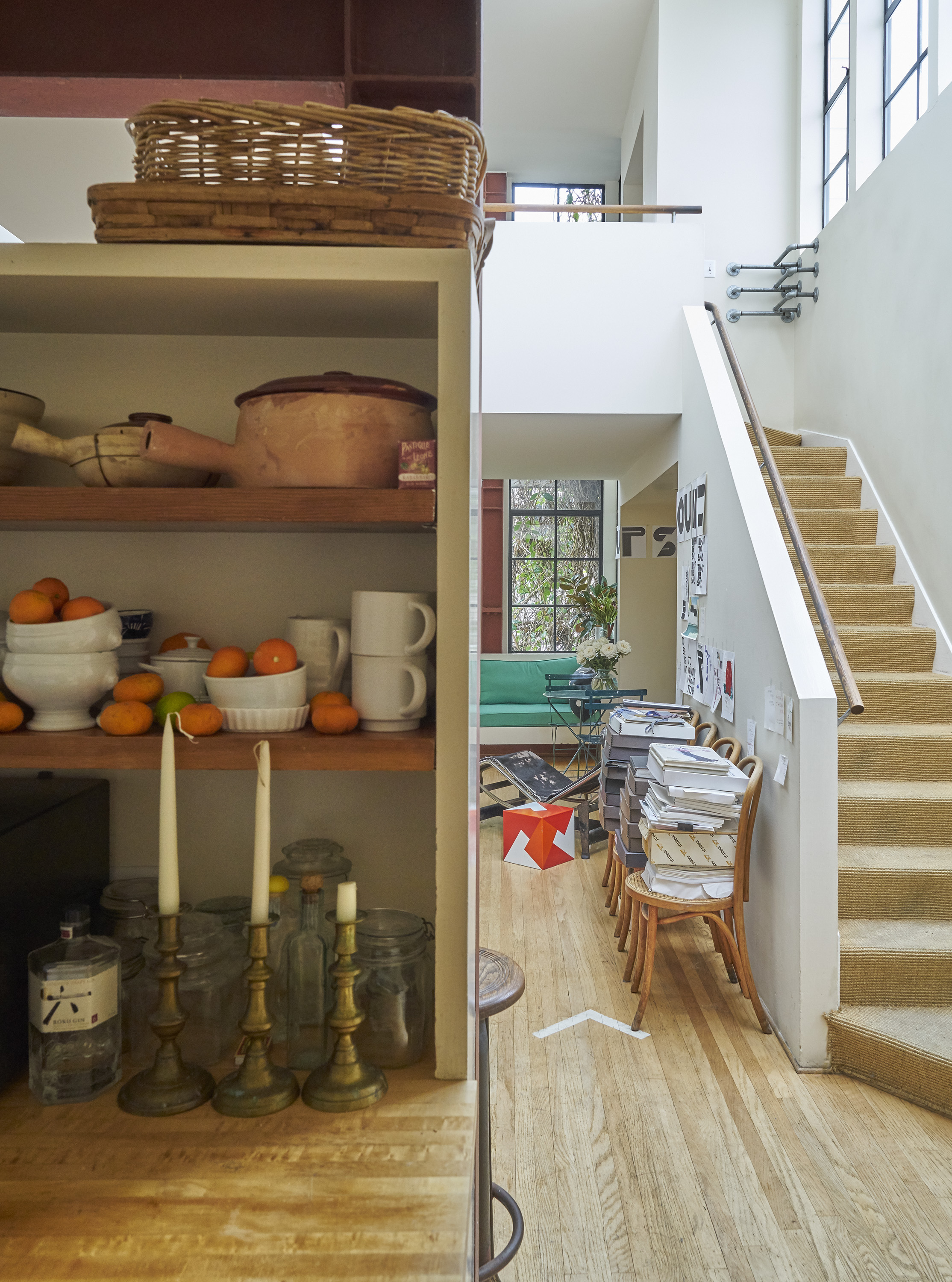 A cozy, eclectic home corner with a shelf of fruits and pottery, a staircase, a stack of books, chairs, and glimpses of a green sofa and art supplies.