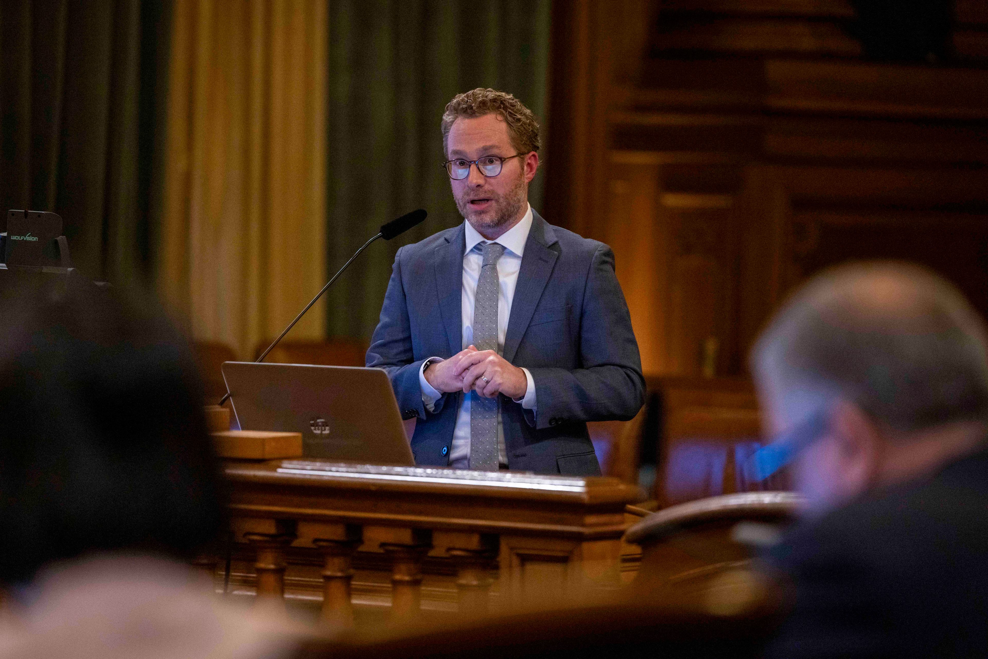 A man stands at a podium speaking, his hands clasped, in an ornate wooden room. He's wearing a gray suit.