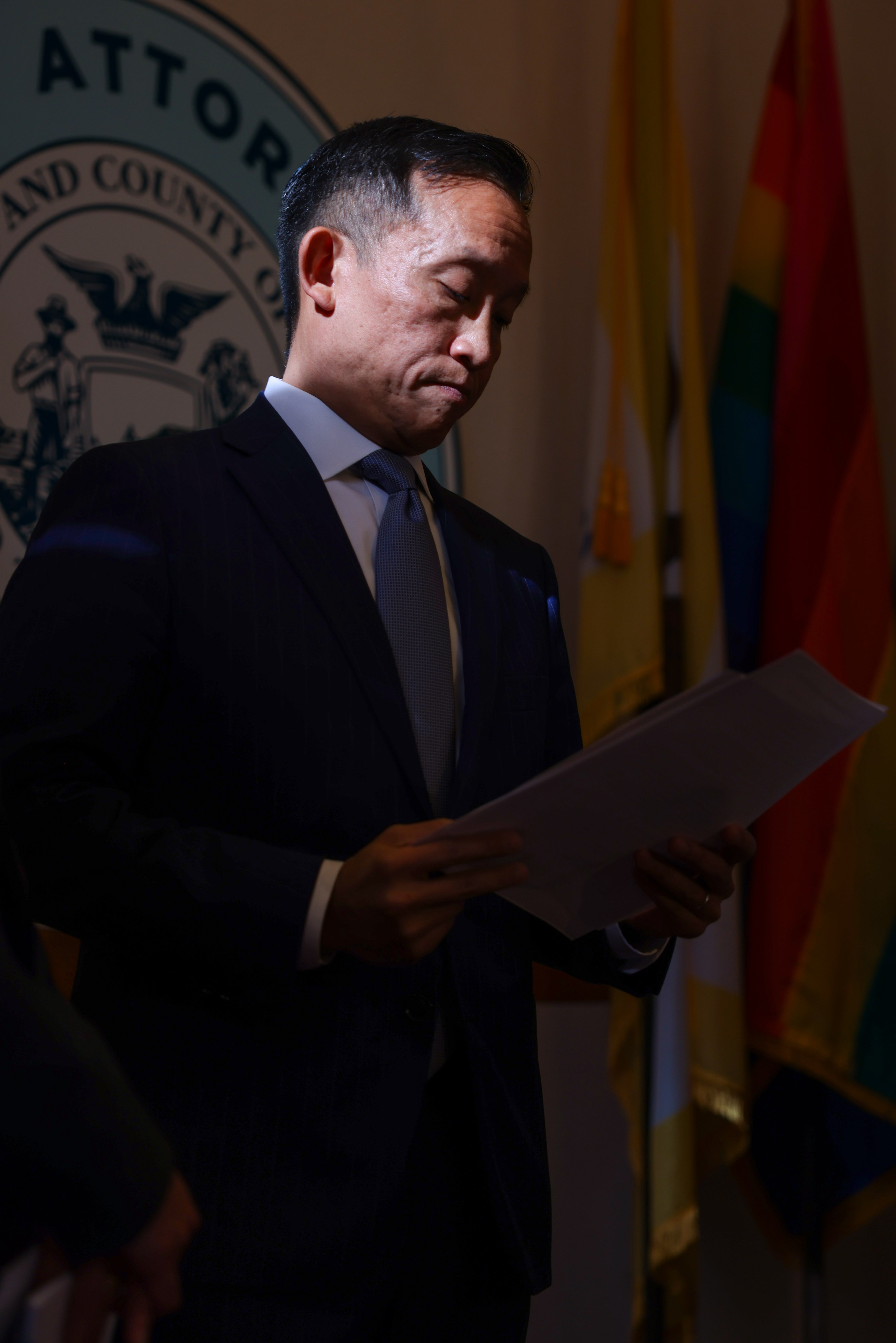 A man in a suit stands in dim light, reading papers. Behind him are flags and a seal with an official emblem partially visible.