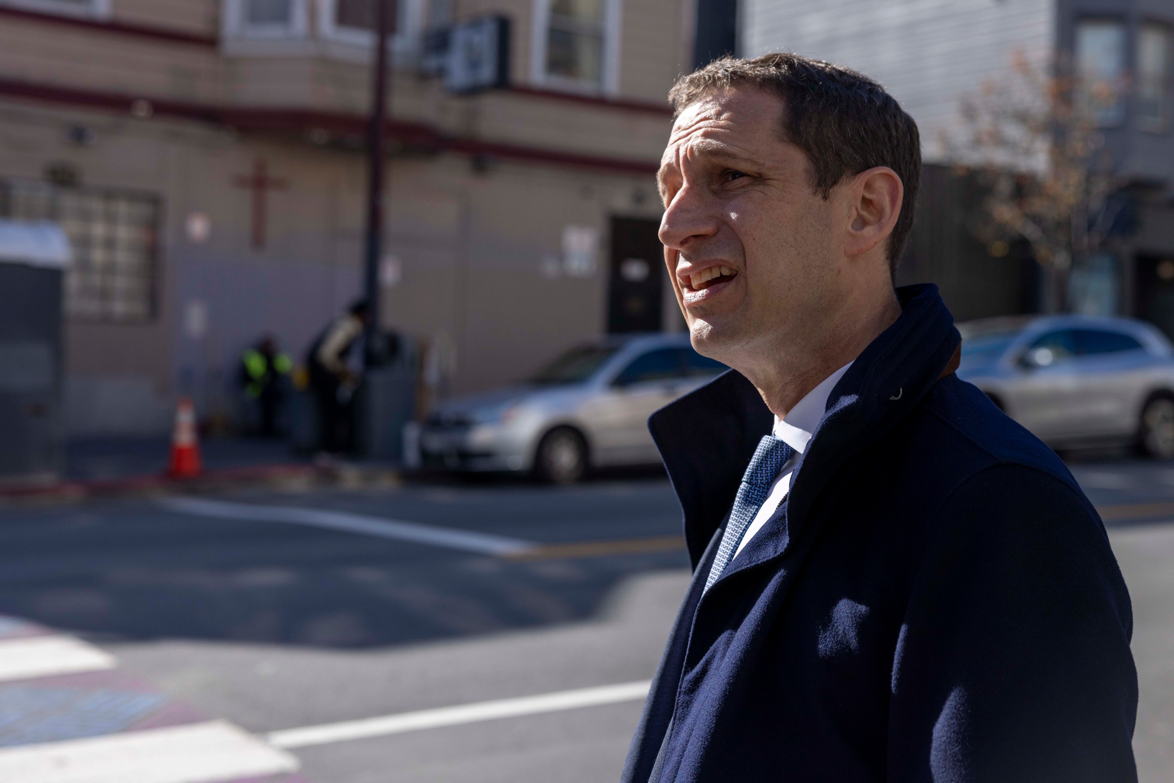 A man in a dark coat stands on a sunny street, speaking. In the background, there's a building with a cross, a car, and people near a traffic cone.