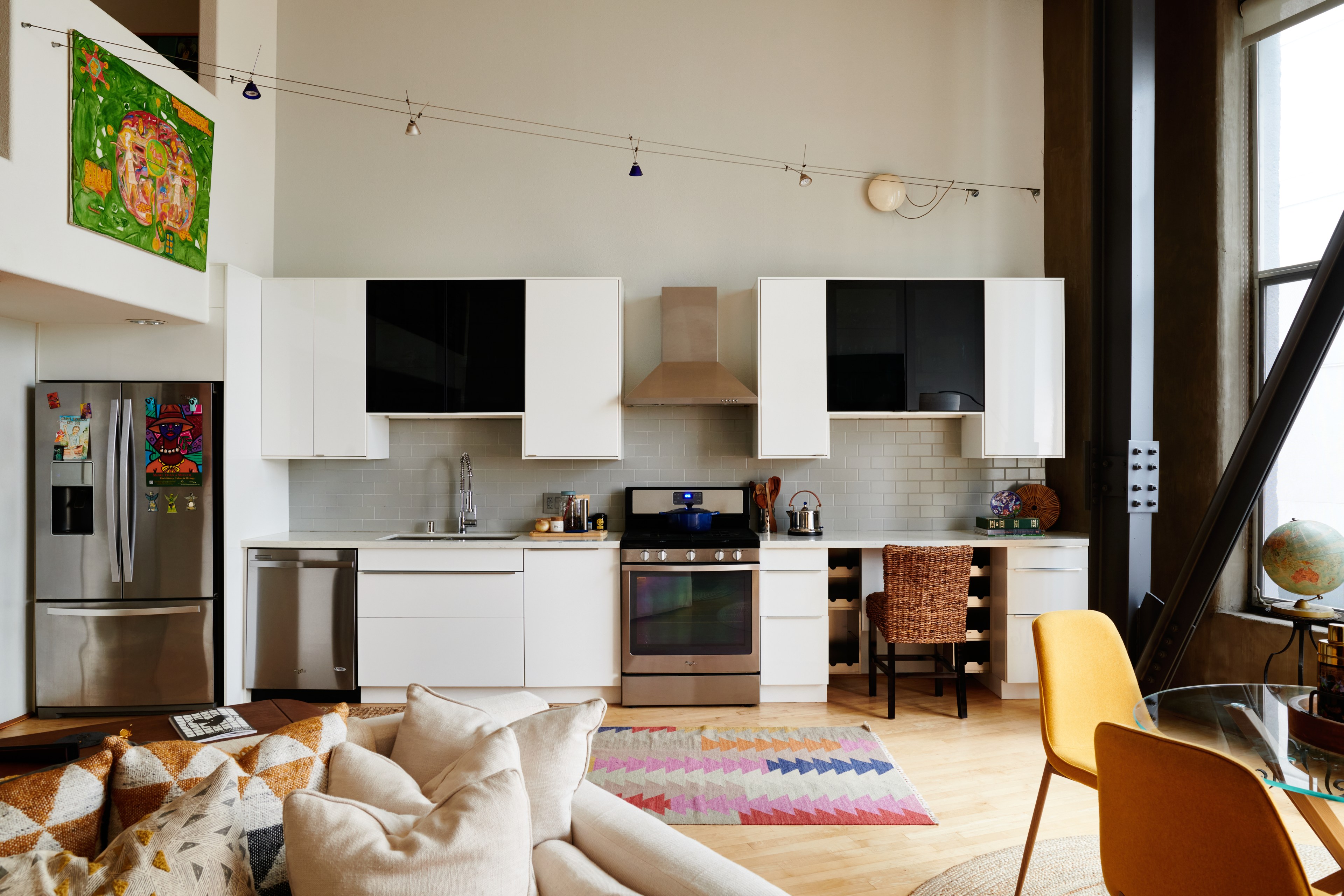 A modern kitchen with white cabinets, stainless steel appliances, a colorful rug, and art on the wall. There's a seating area with a couch and a glass table.