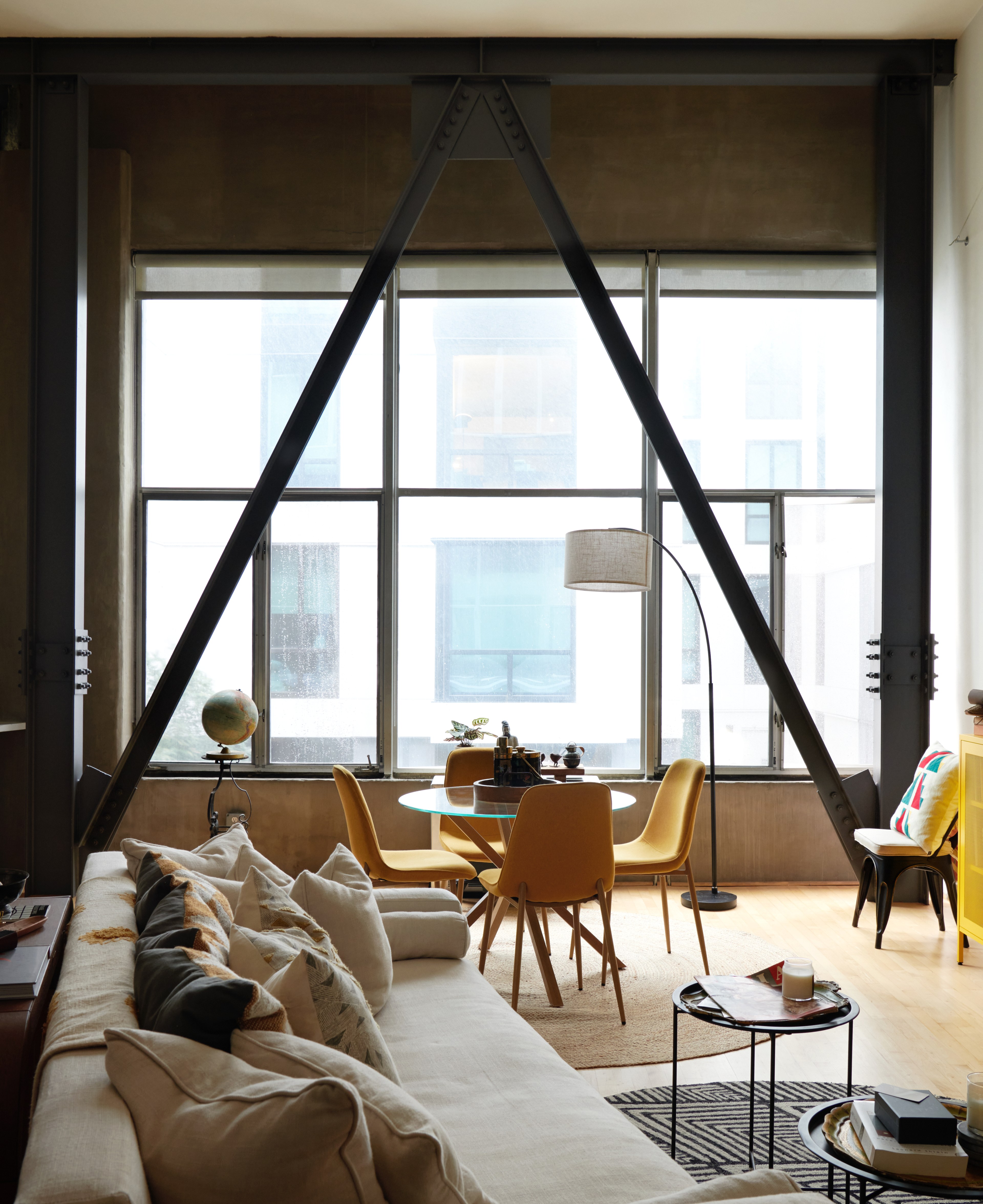 A modern living room features a large window, a beige sofa with cushions, a round glass table with yellow chairs, an arc lamp, and a globe near the corner.