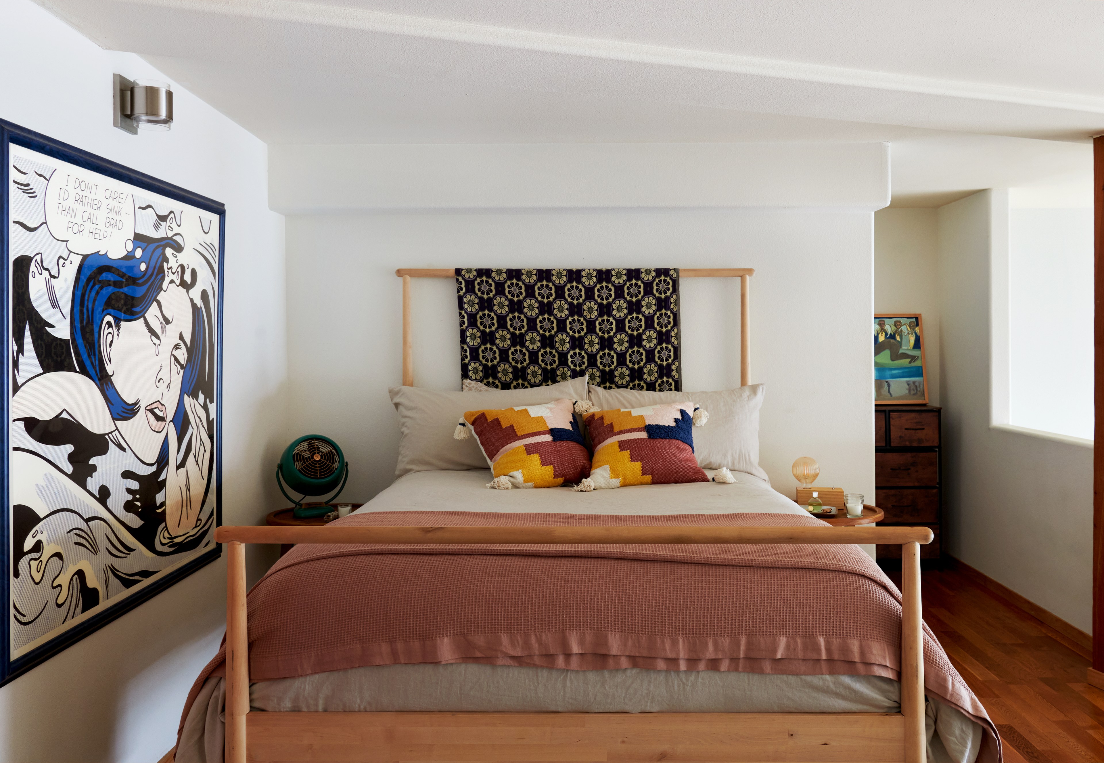 A room features a wooden bed with colorful pillows and a blanket. A pop art painting is on the wall, and there's a small fan on the bedside table.