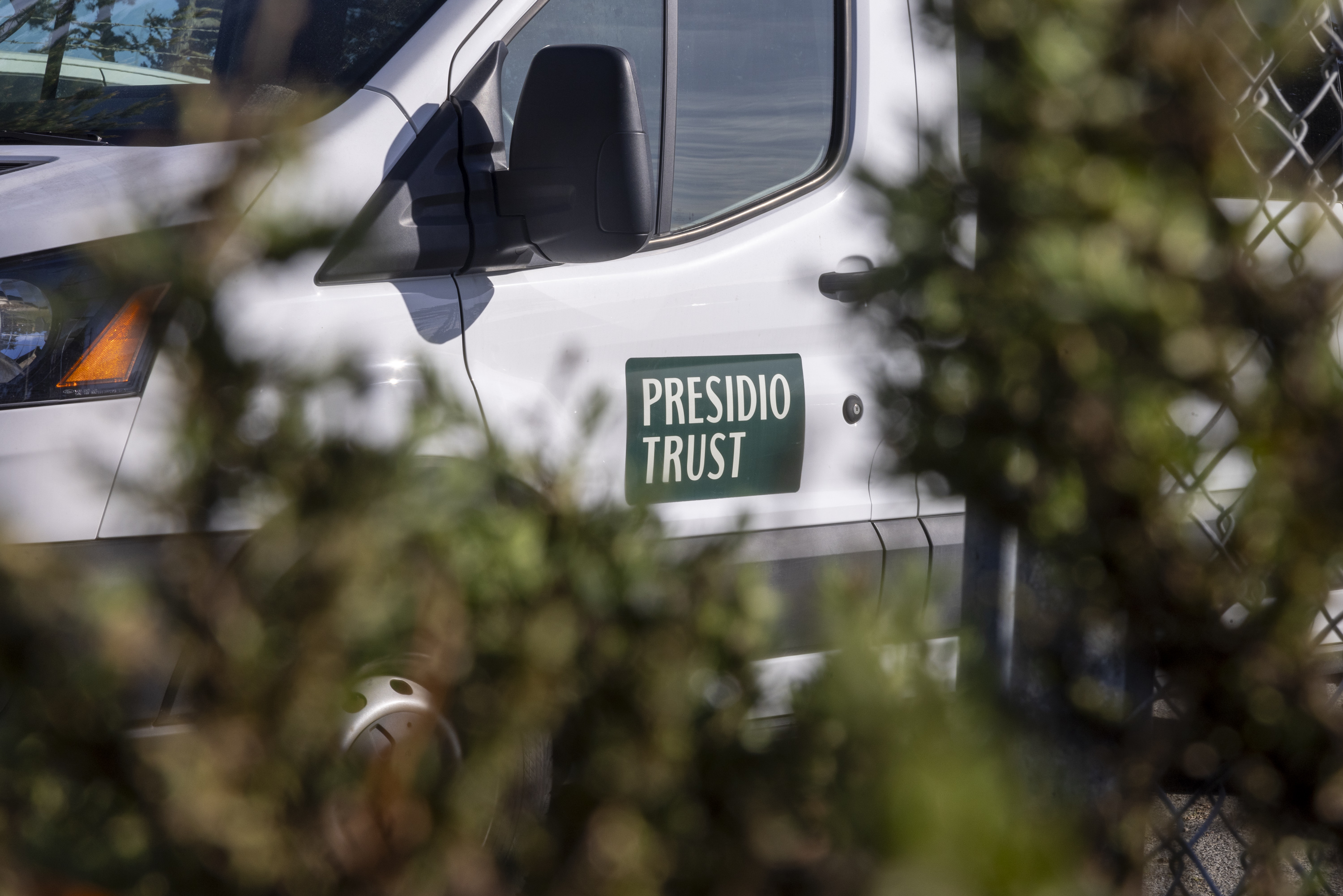 A white vehicle with &quot;Presidio Trust&quot; written on the side is partly visible behind blurred greenery, with a chain-link fence in the background.