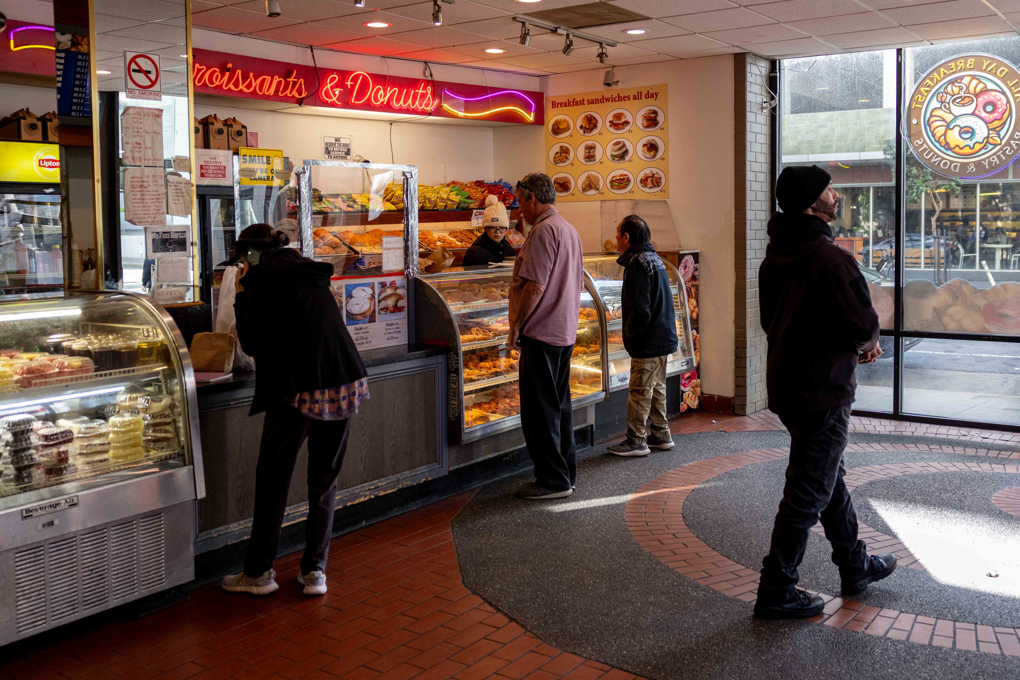 Customers are inside a bakery shop with a neon "Croissants & Donuts" sign, ordering from a glass display filled with pastries. A large window is on the right.