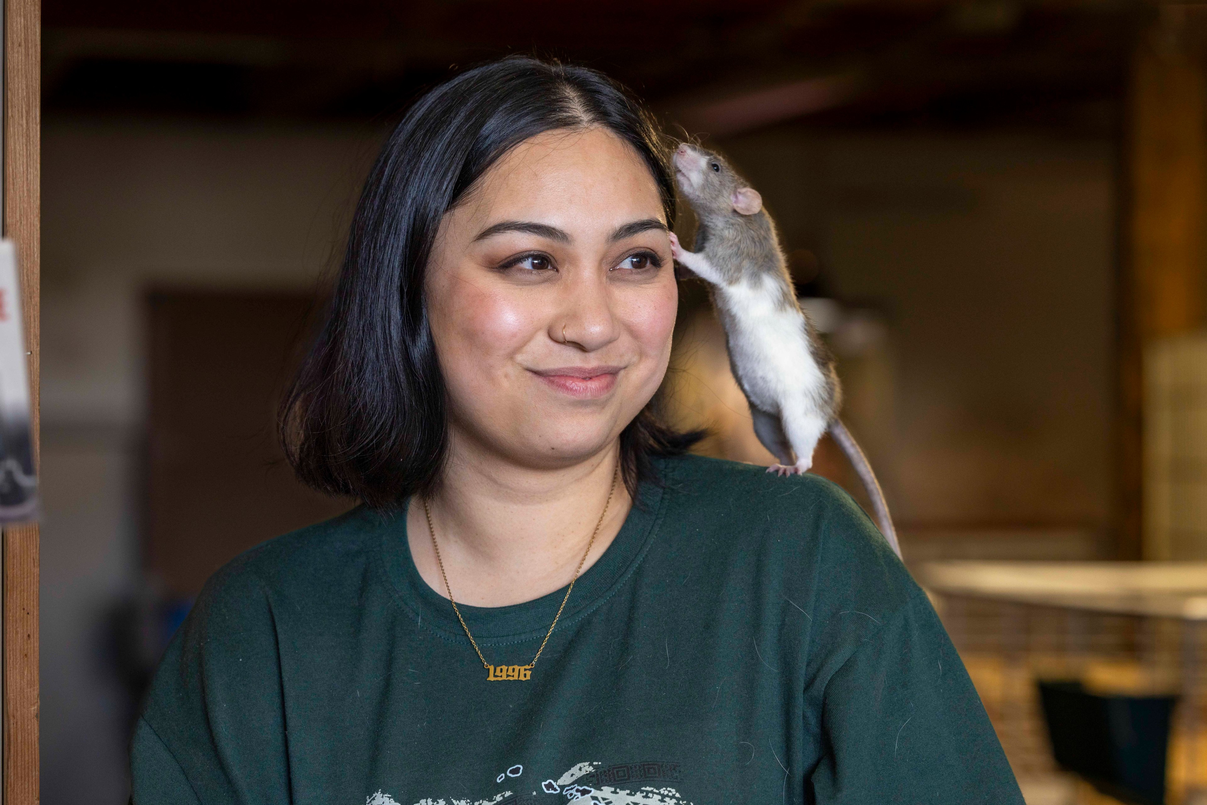 A woman with shoulder-length dark hair smiles as a rat perches on her shoulder, sniffing near her ear. She wears a green shirt and a necklace with "1996."