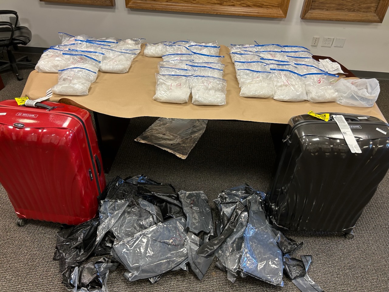 The image shows a table with numerous clear plastic bags containing white substances, flanked by a red and a black suitcase. Black plastic wraps are on the floor.