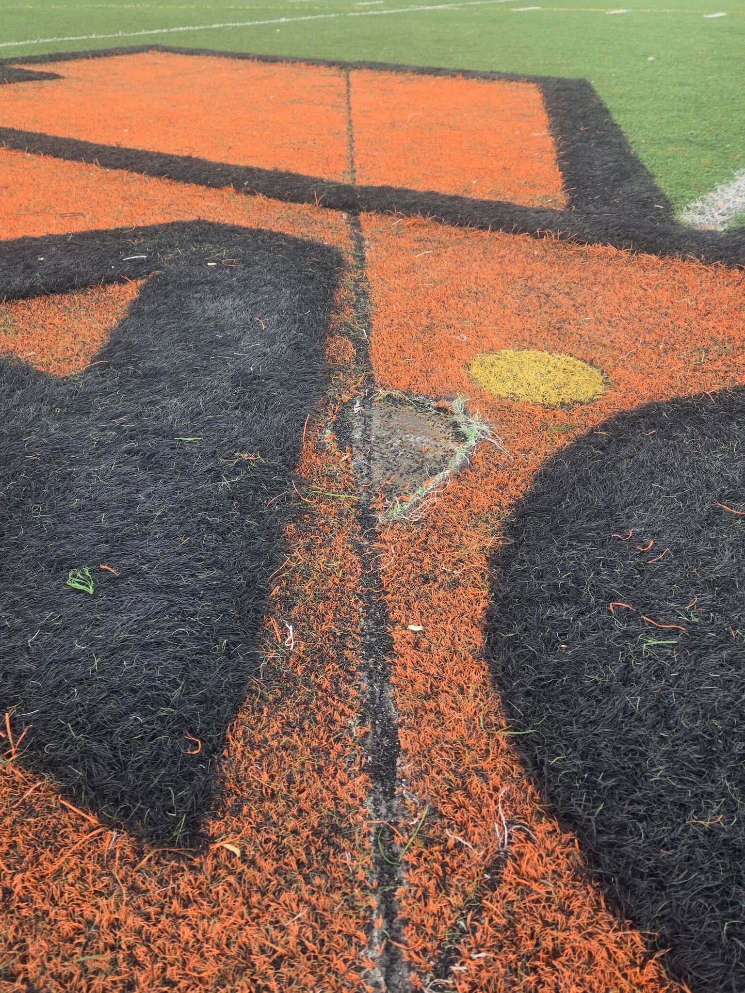 The image shows a section of artificial turf with a design featuring orange and black shapes, including a yellow circle, and a noticeable worn patch in the center.