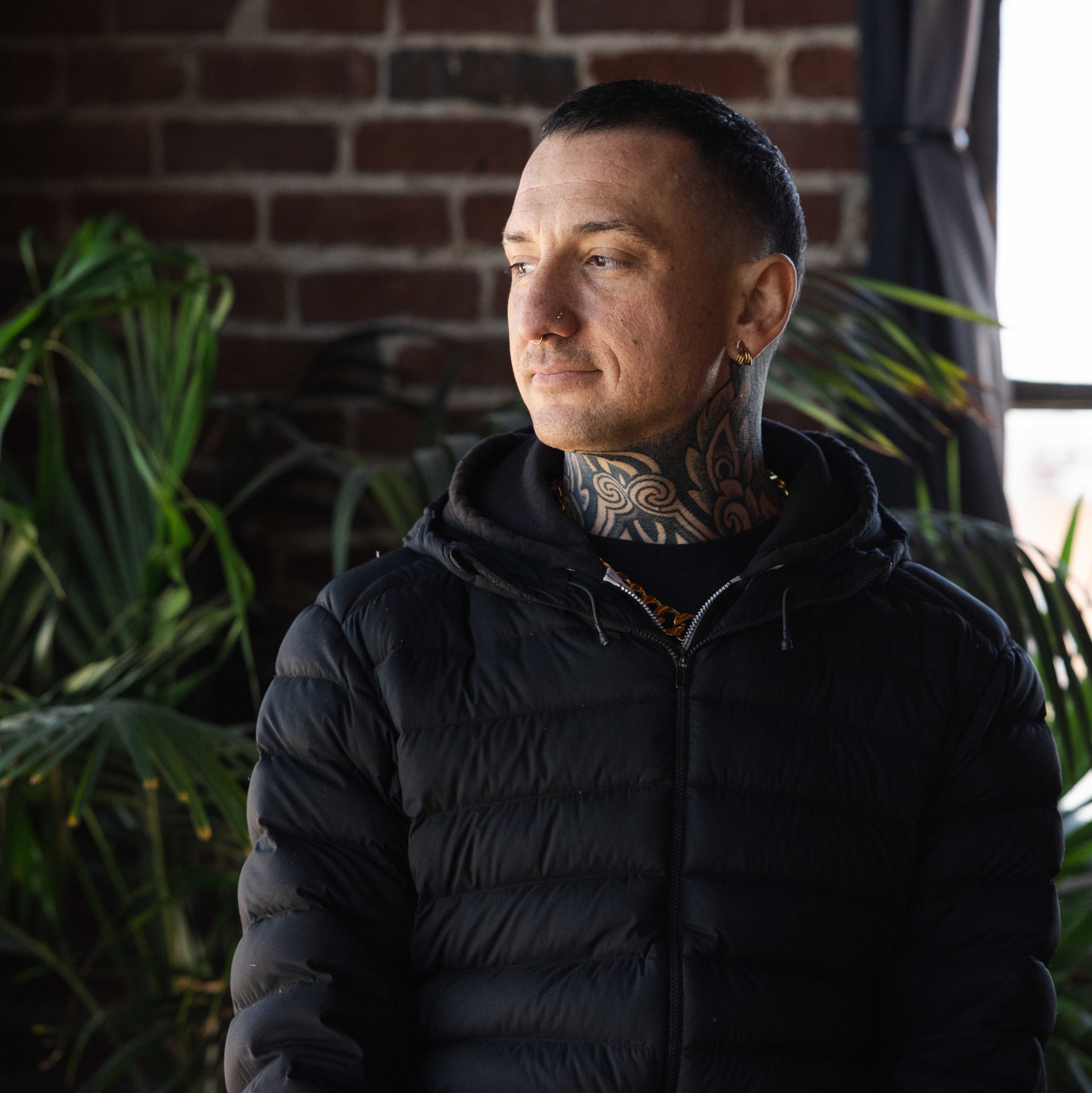 A person with a tattooed neck, wearing a black puffer jacket, looks thoughtfully to the side. They stand indoors near brick walls and green plants.