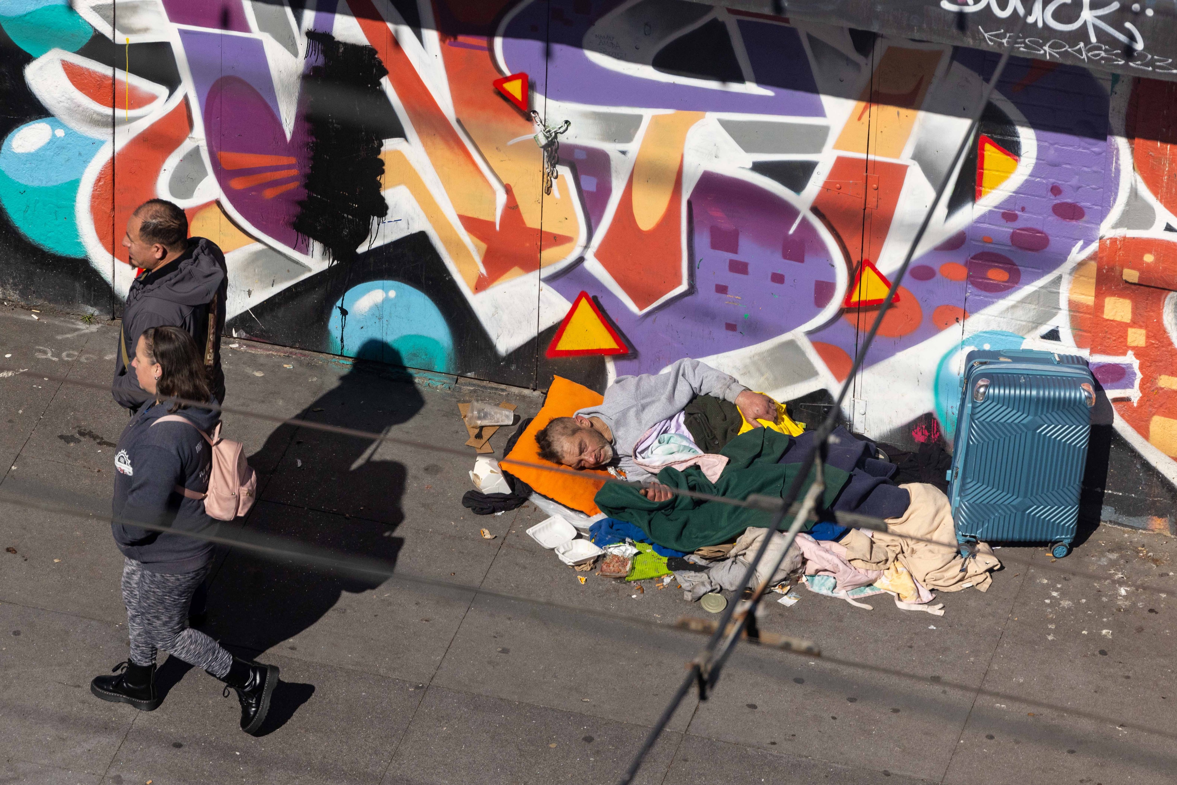 A person sleeps on a sidewalk surrounded by blankets and a suitcase, beside a colorful graffiti wall, while two people walk past.