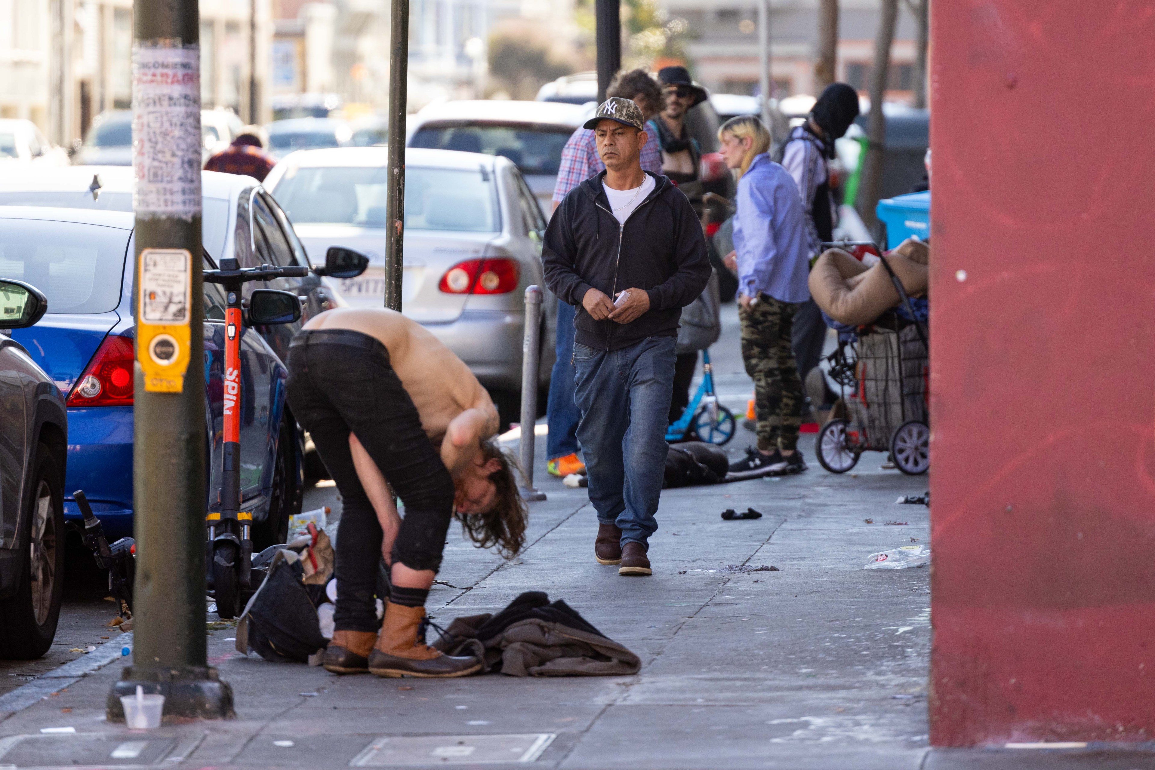 A busy city sidewalk is crowded with people, one bent over with no shirt. Others stand or walk past, and there are parked cars and a scooter nearby.