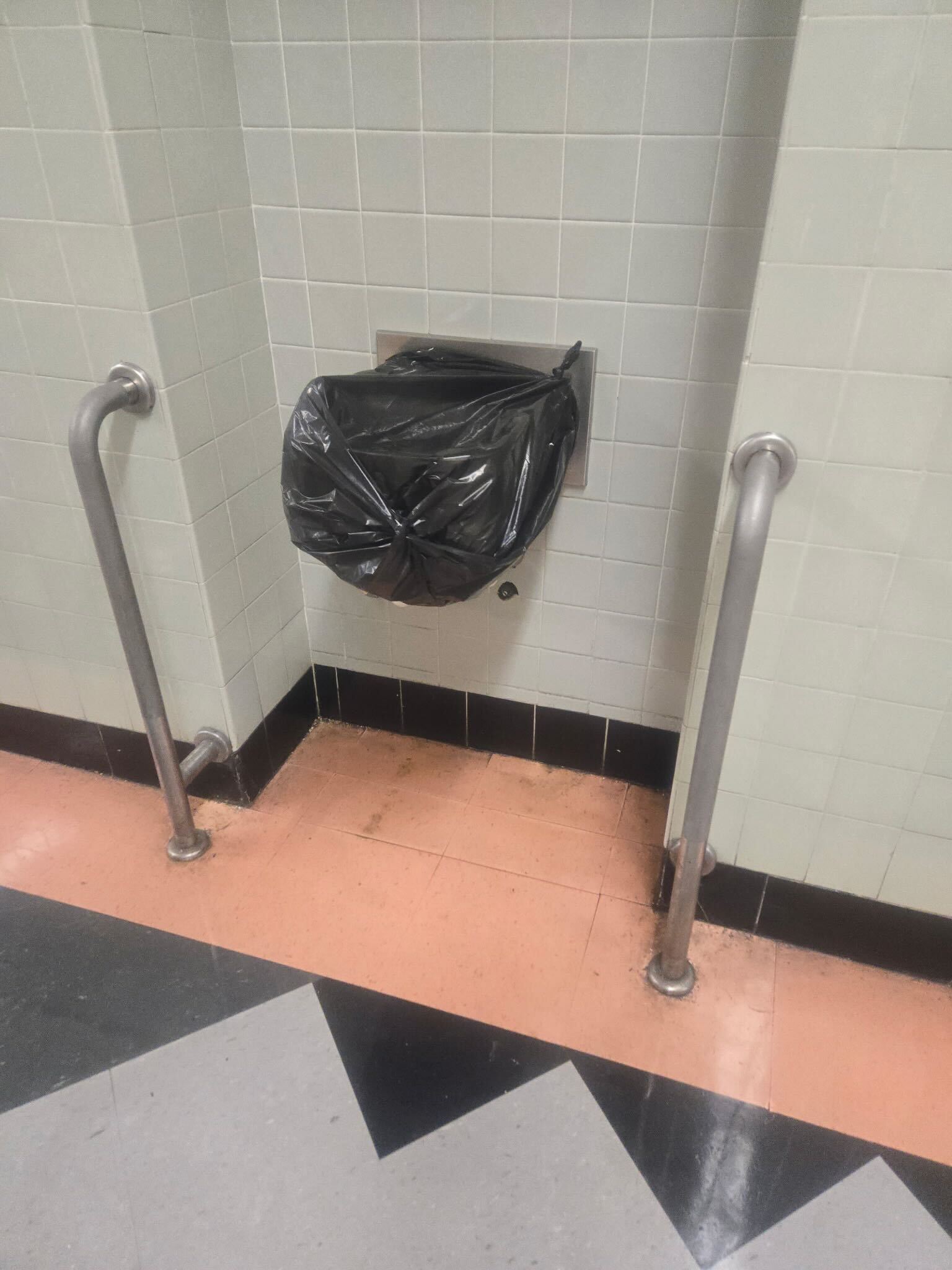 A wall-mounted hand dryer is covered with a black plastic bag, flanked by metal railings on both sides. The floor has peach, black, and gray tile patterns.