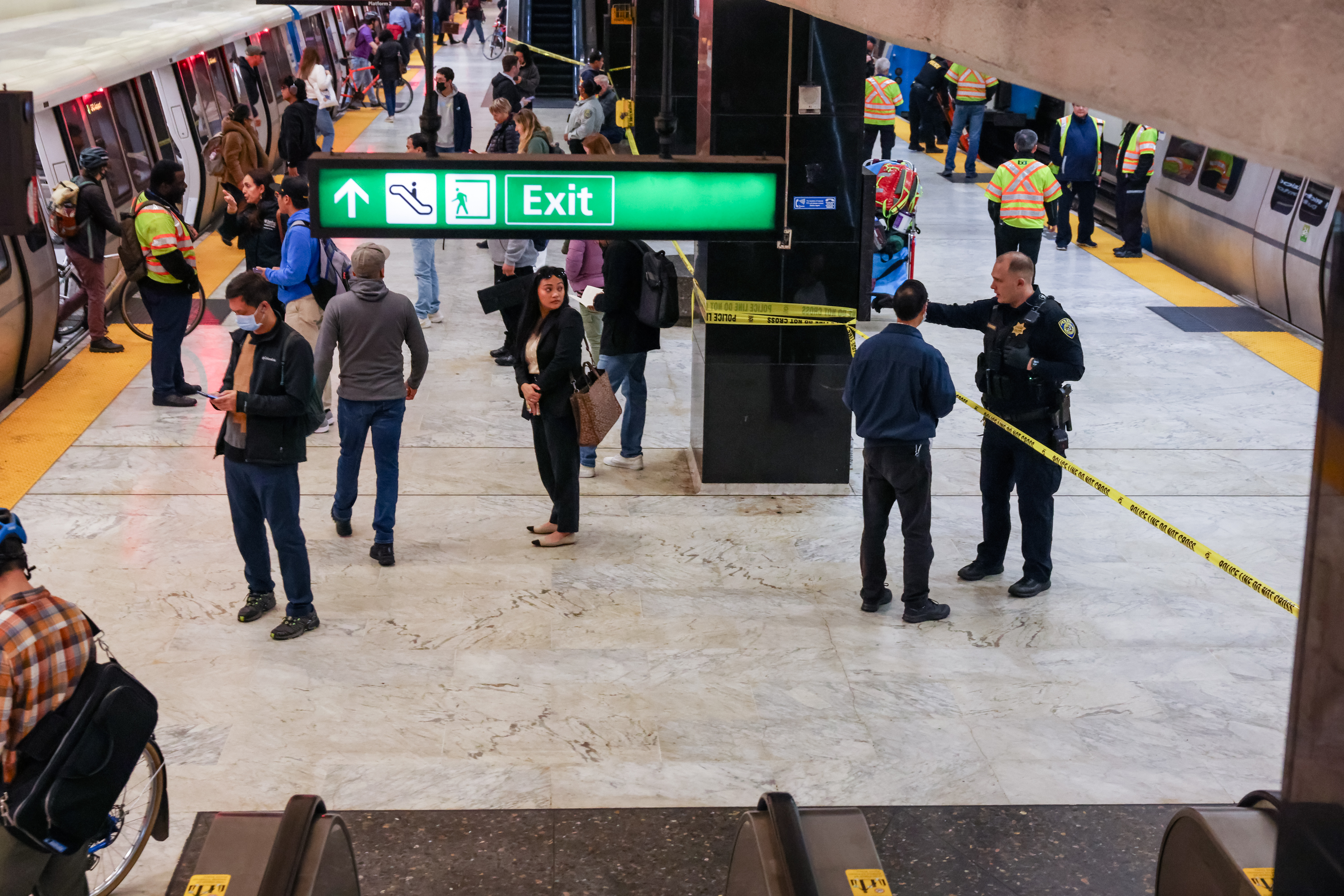 A busy subway station features a train on the left, a green exit sign above, several people standing around, and police tape marking off an area, with officials present.