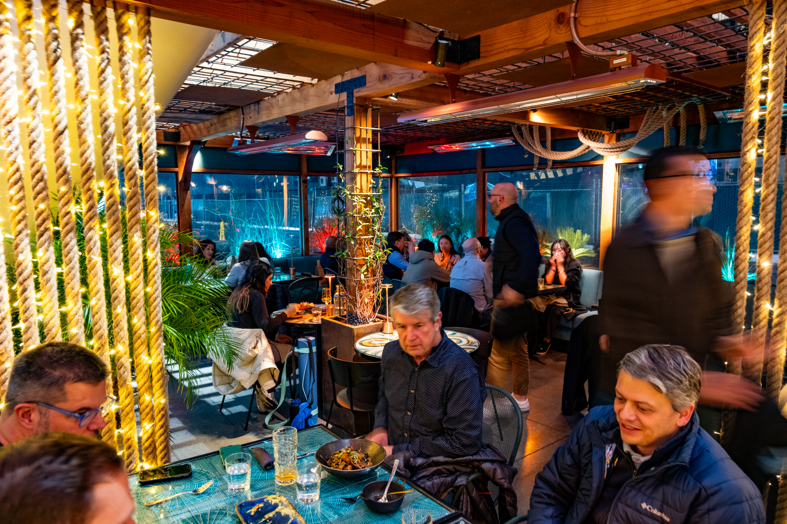 A warmly lit restaurant with people dining at tables. Decor includes ropes with fairy lights and plants, creating a cozy atmosphere. Some patrons are blurred in motion.